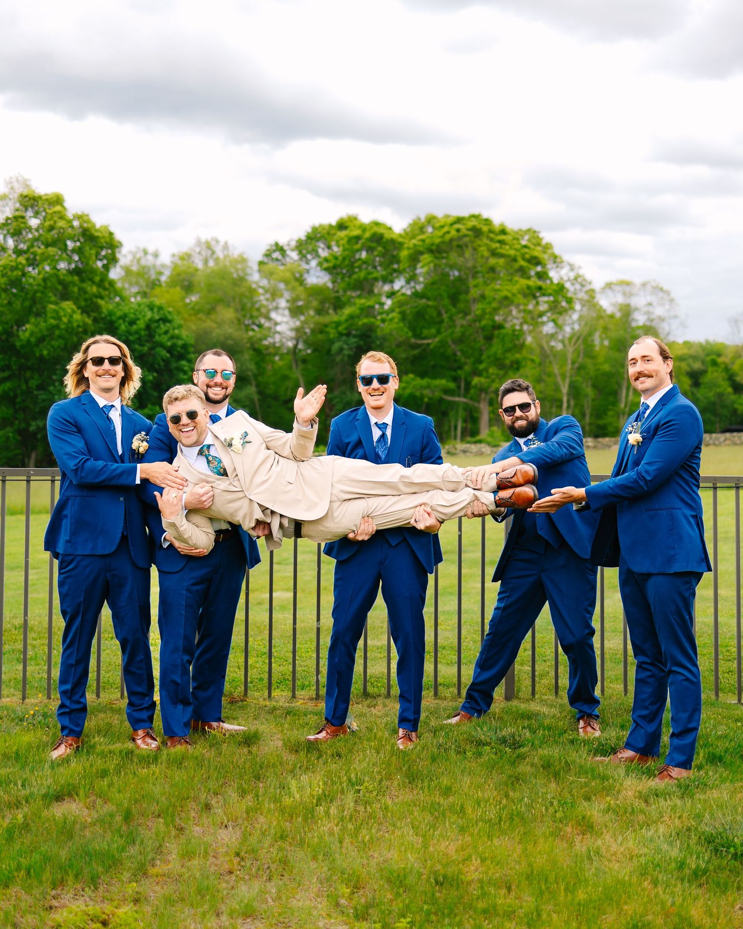 Groomsmen wearing matching navy blue suits posing playfully on a grassy field with trees in the background in New London County Connecticut