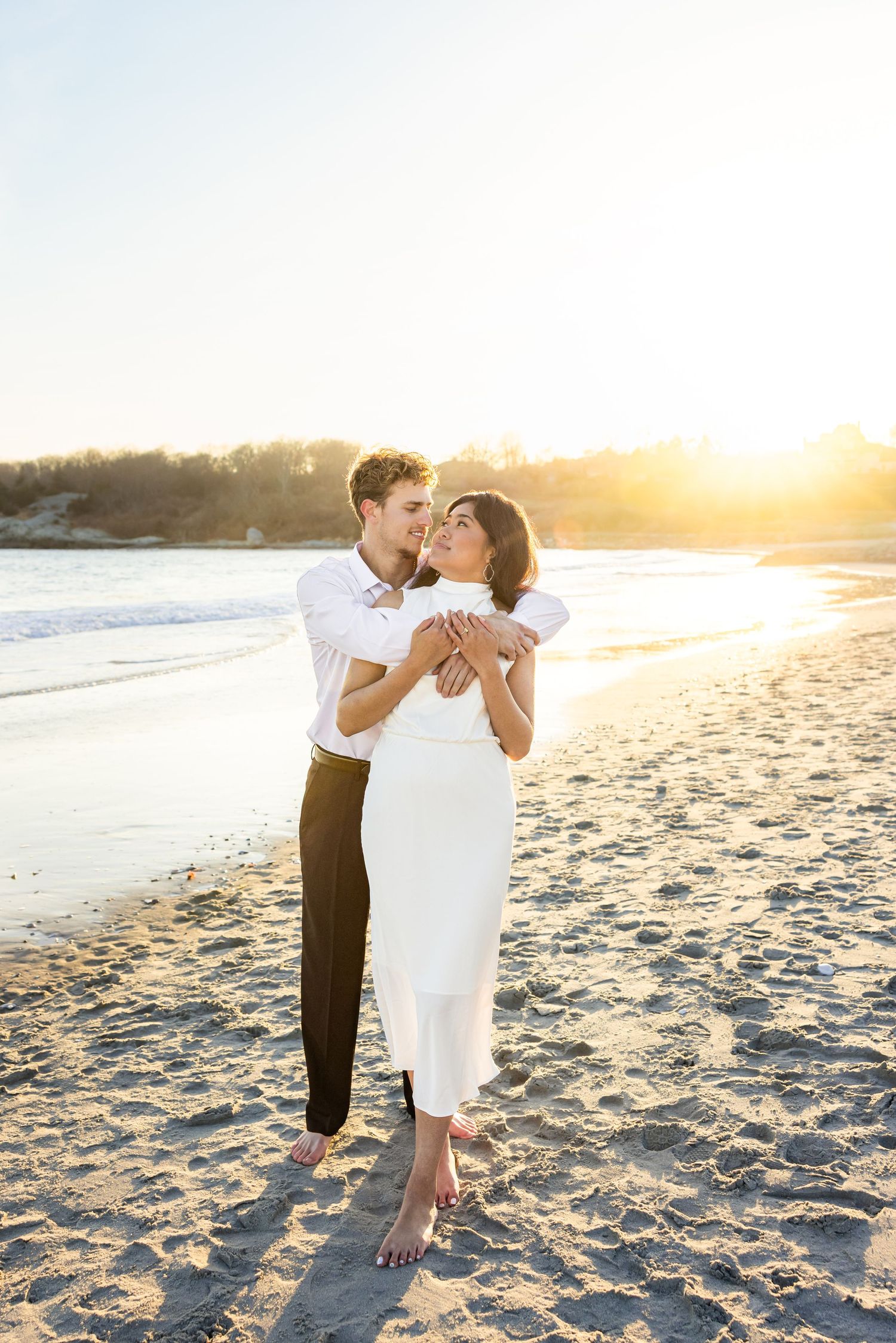 A couple embraces on a sunny beach at golden hour as waves crash on the shore in the background.