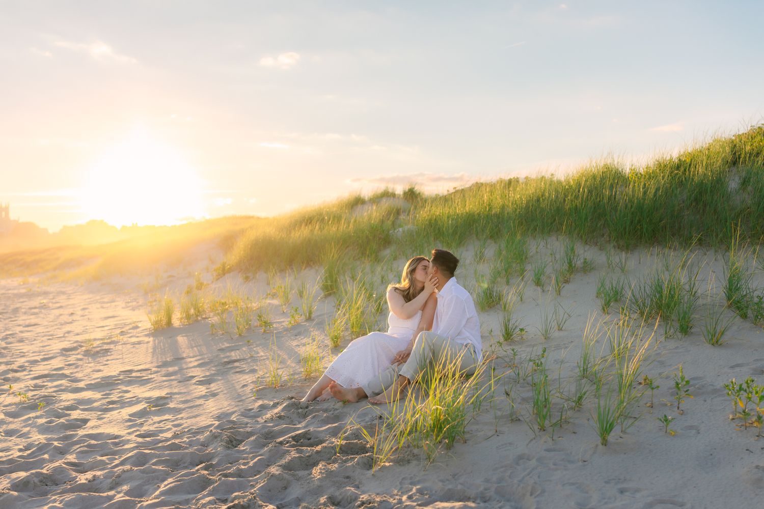 Romantic couple sitting together on sandy beach dunes during a golden sunset, both dressed in white.