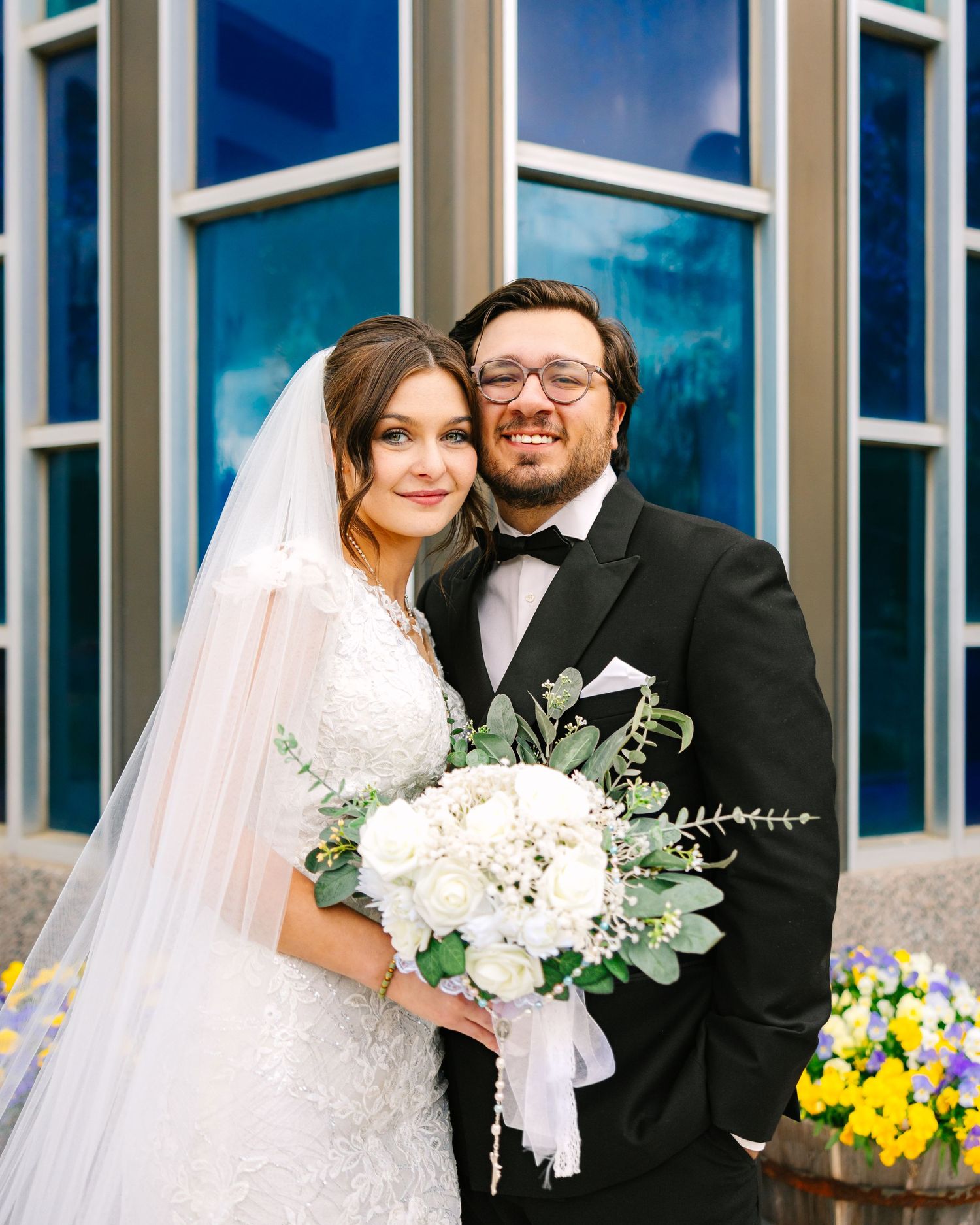 Newlywed couple posing outside church, bride in lace gown holding white bouquet, groom in black tuxedo.