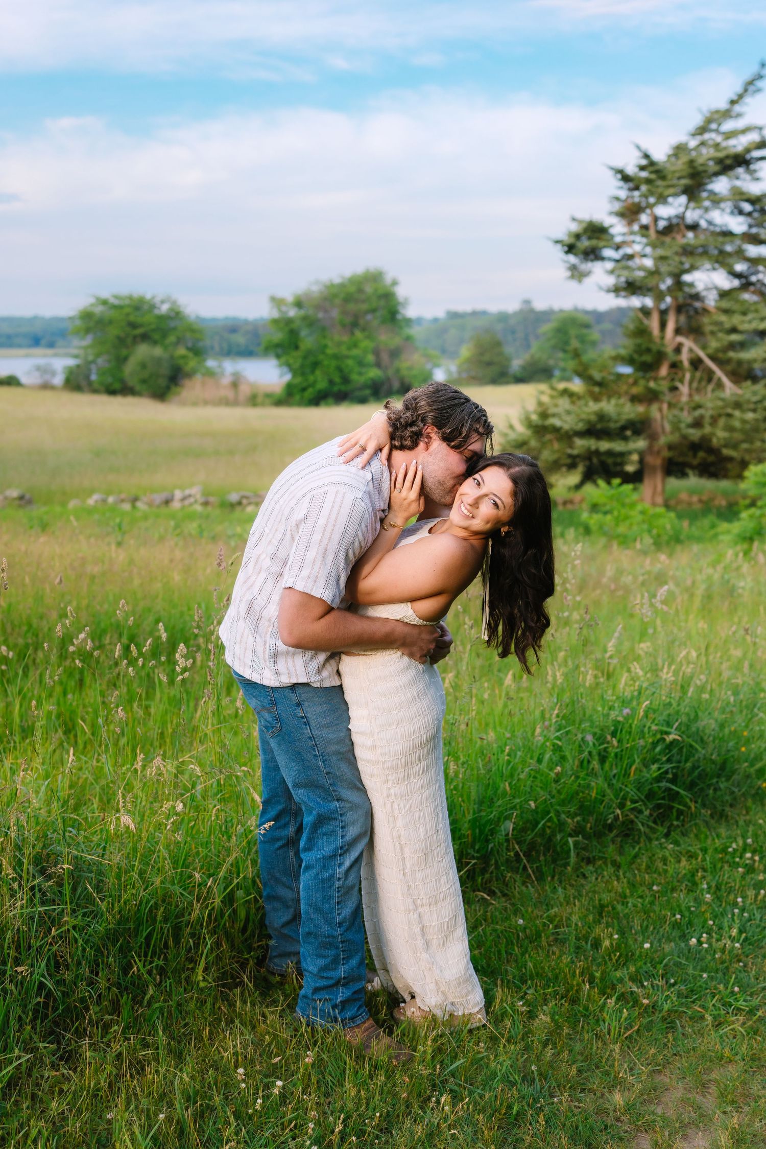 Man kissing woman's cheek as she smiles in a green field with a lake in the background.