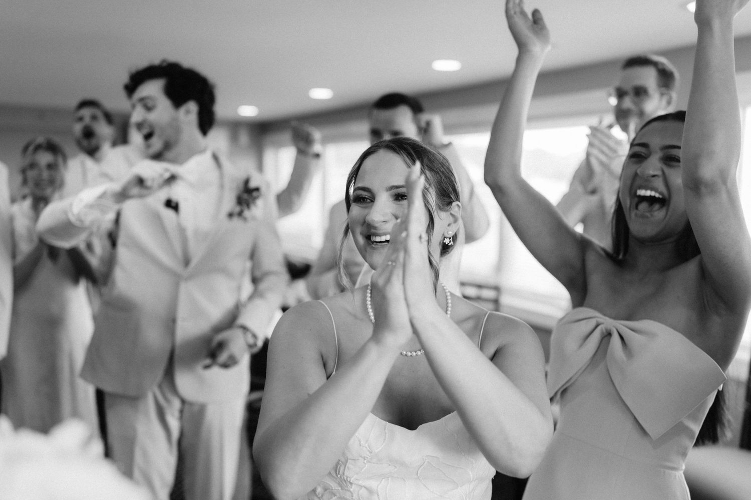 Black and white photo of joyful bride clapping with hands together surrounded by cheering wedding party guests indoors.