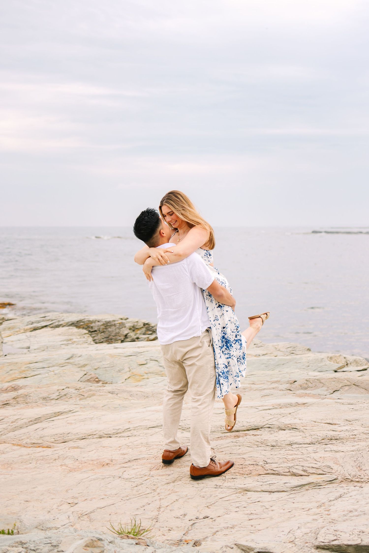 A romantic couple embracing and dancing together on rocky coastal shores at sunset after surprise proposal on Ocean Drive in Newport RI