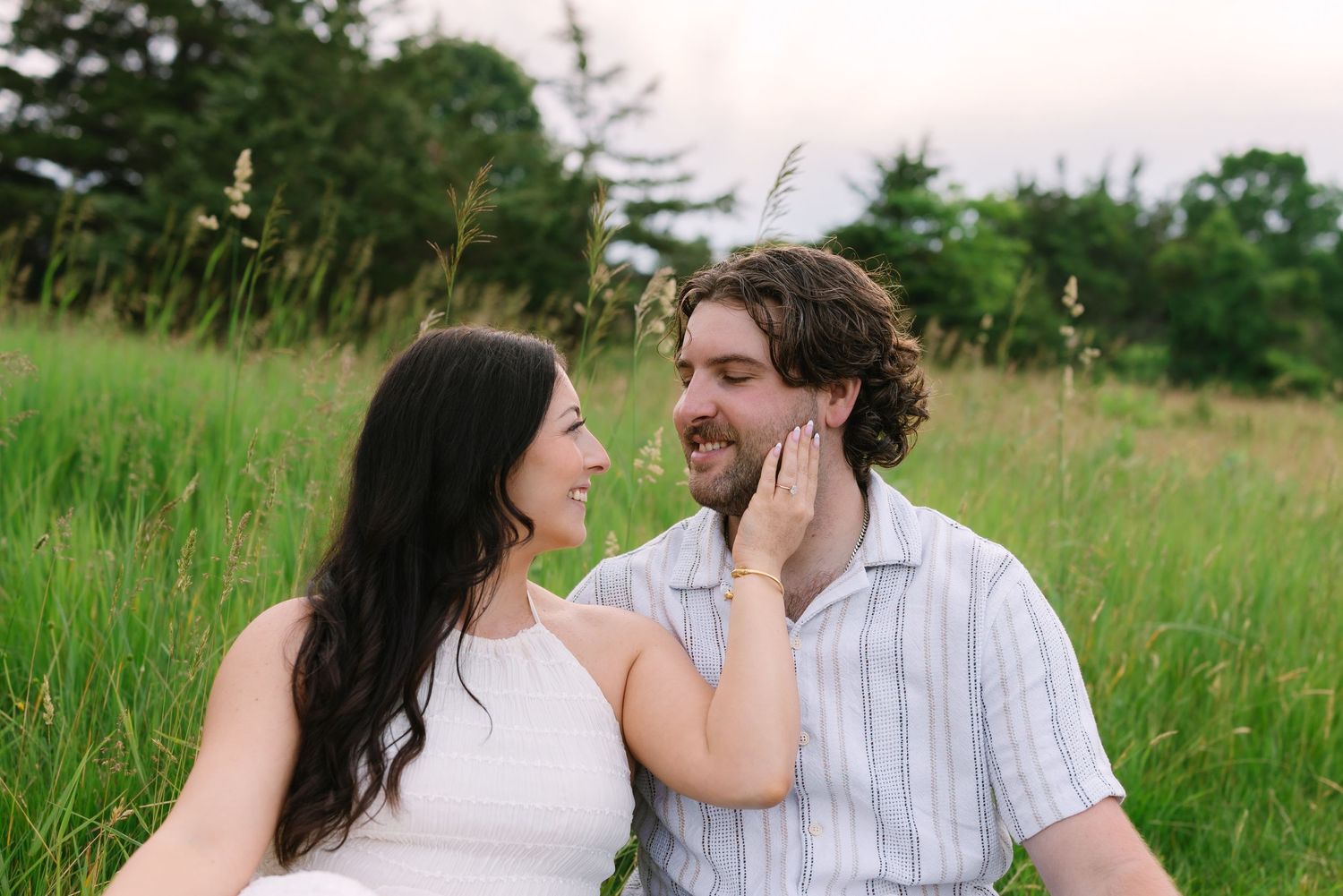 Happy engaged couple sitting in a green field smiling at each other at sunset.