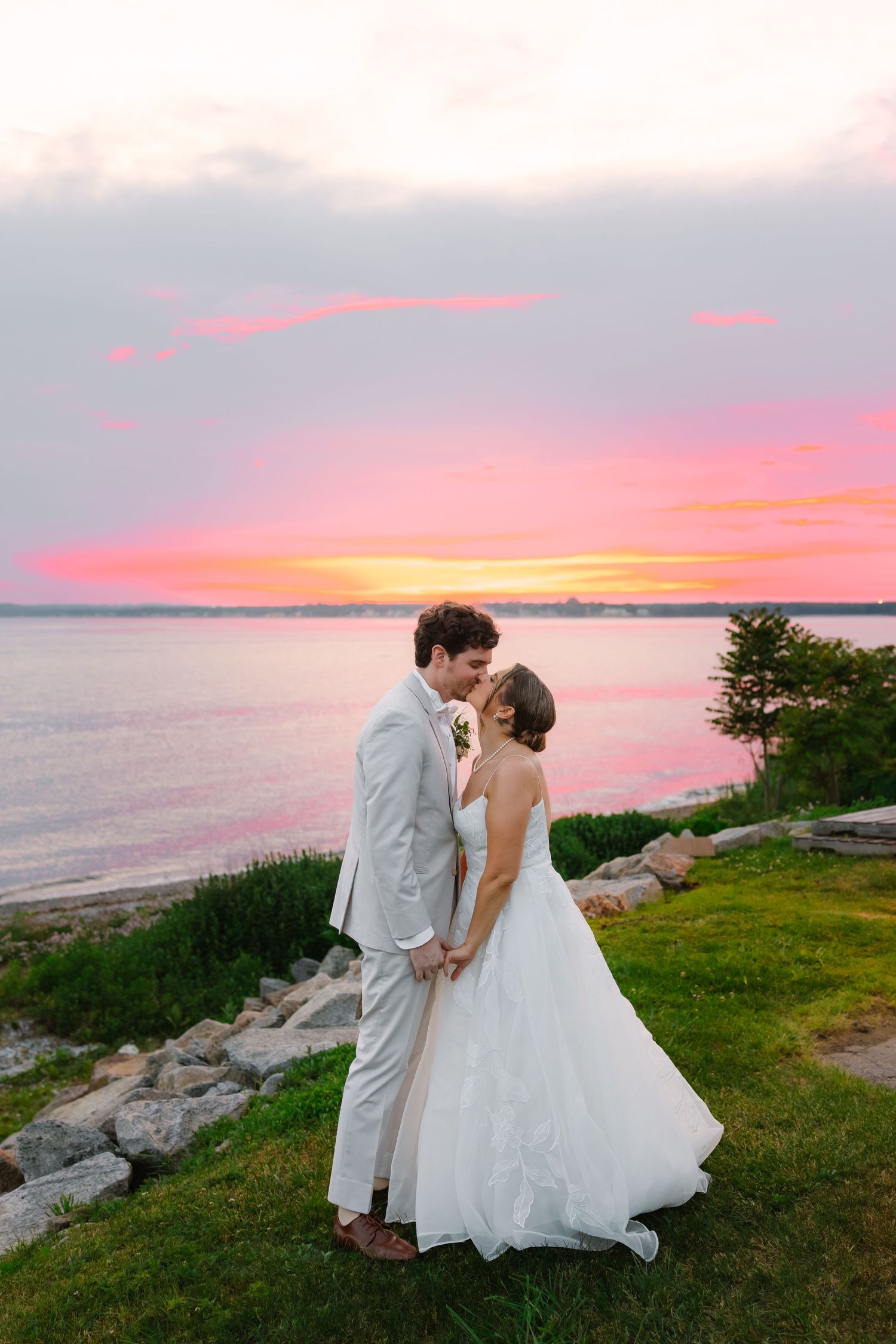 Bride and groom sharing a kiss on grassy waterfront during a vibrant pink and orange sunset.