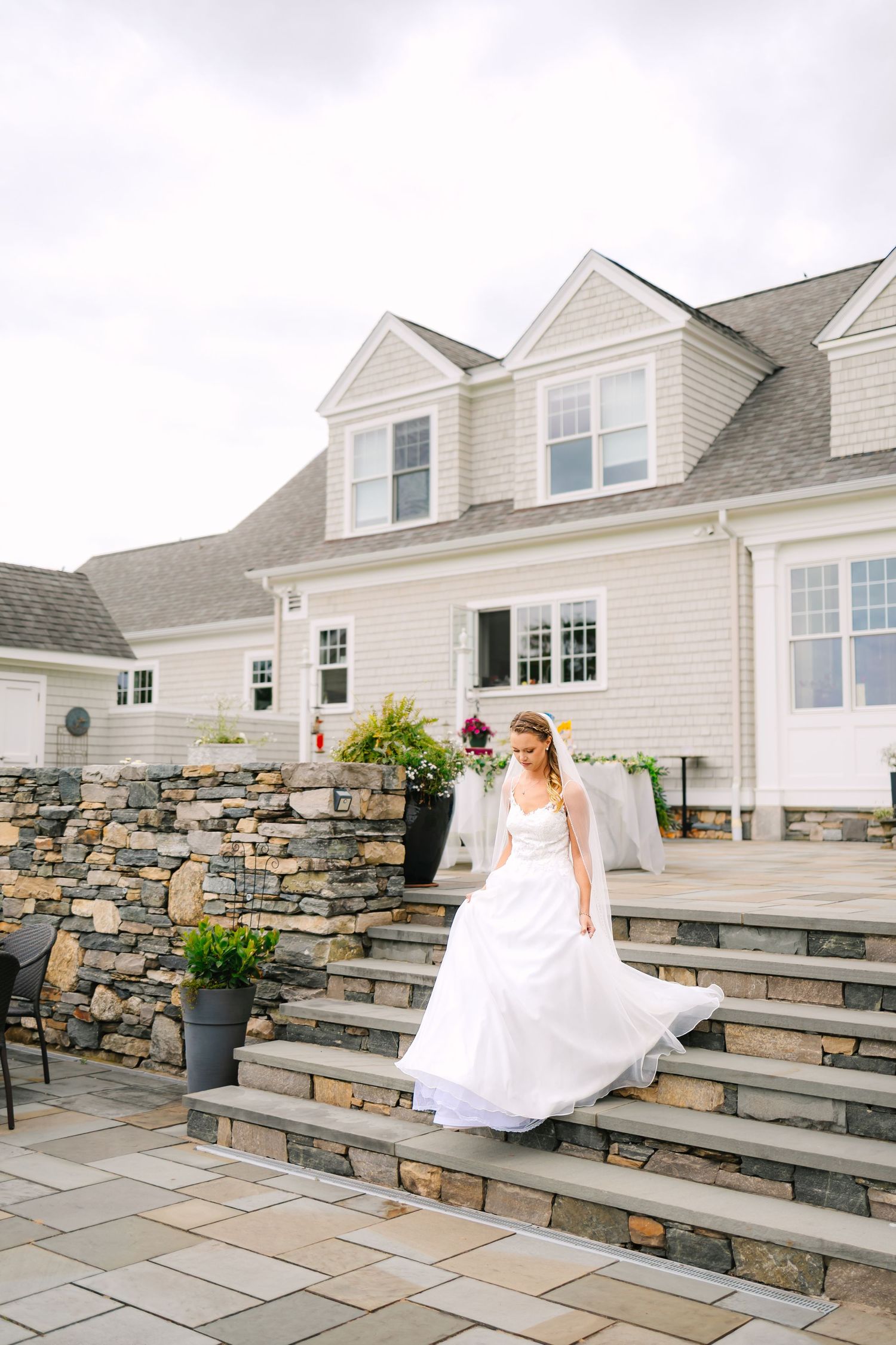 A white wedding dress flows elegantly down stone steps in front of a classic New England style home with gray stonework.