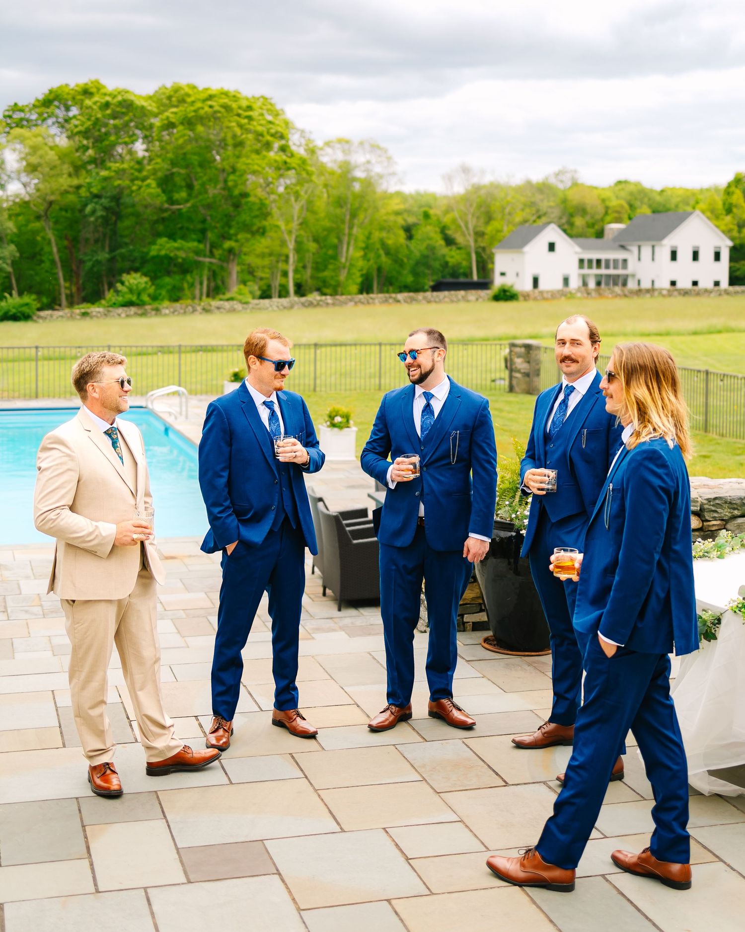 Wedding party in blue suits and one in beige gather on a stone patio near a swimming pool with white house in background.