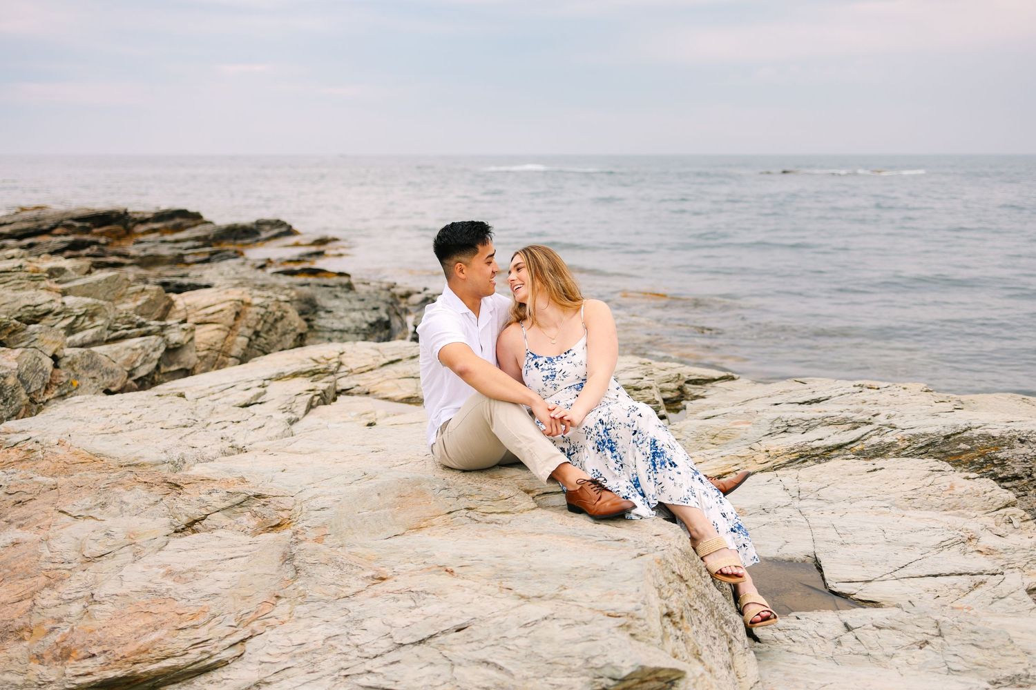 Couple sitting on rocky coastal shore for engagement photos, sharing a romantic moment with ocean waves on Ocean Drive in Newport RI