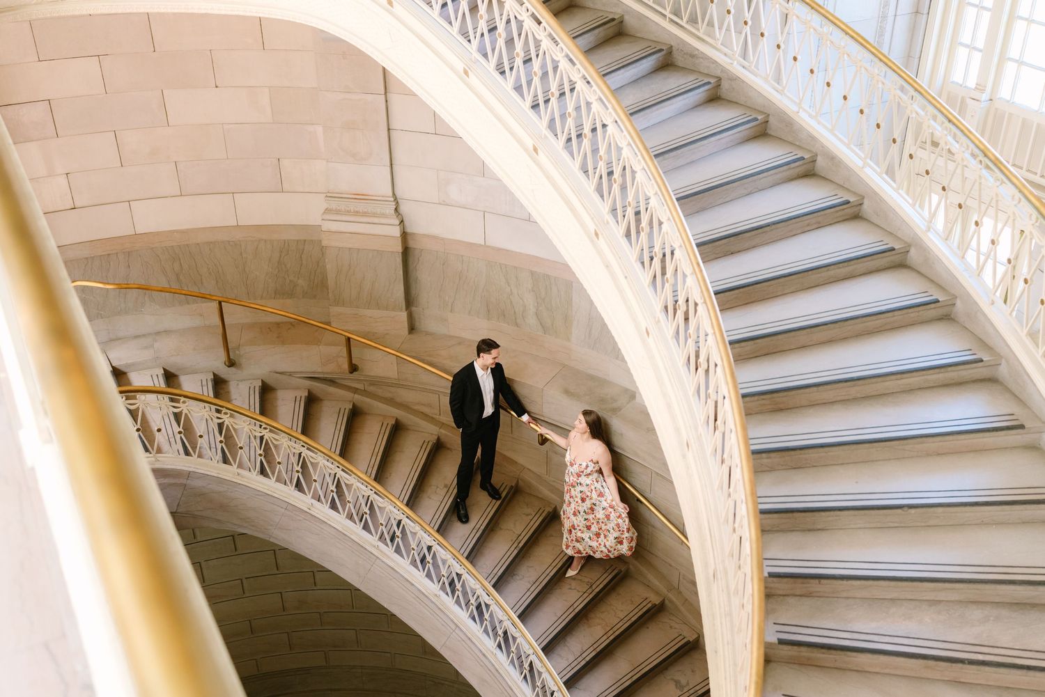 Aerial view of couple holding hands descending elegant curved staircase with ornate white iron railings.