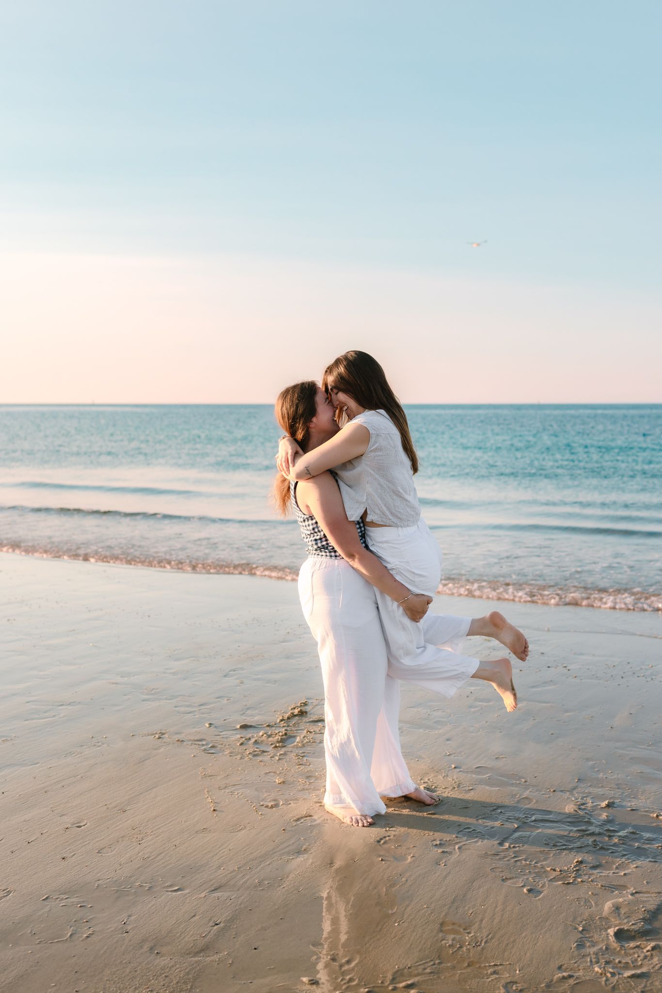 Two women embracing and kissing on sandy beach at sunset near ocean waves.
