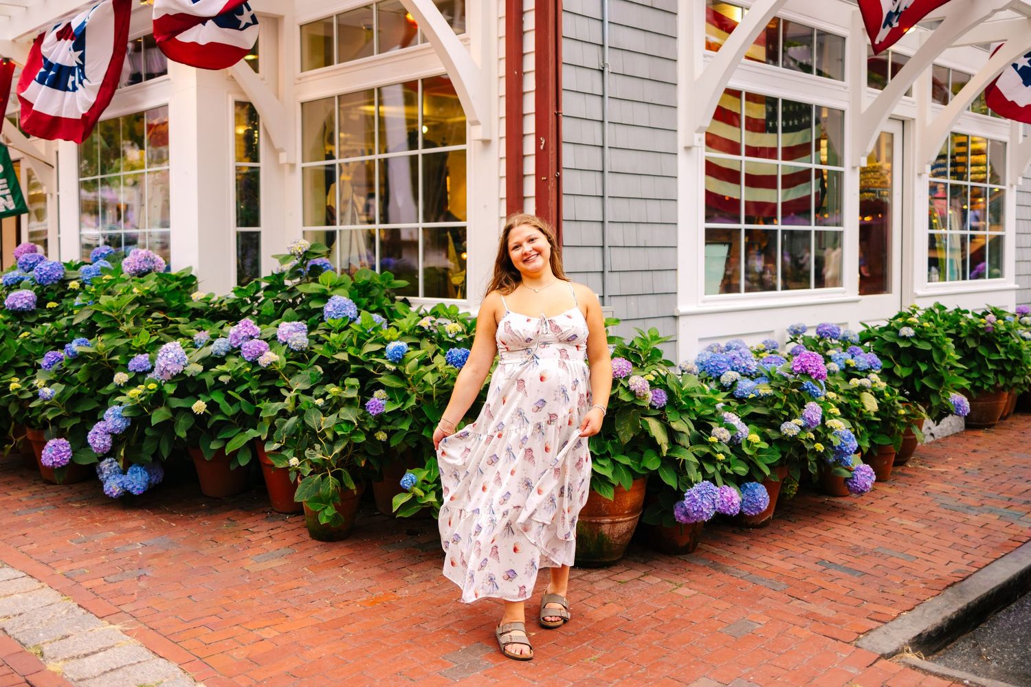 Young woman in floral dress standing before blooming hydrangeas outside a Newport shop with patriotic bunting.