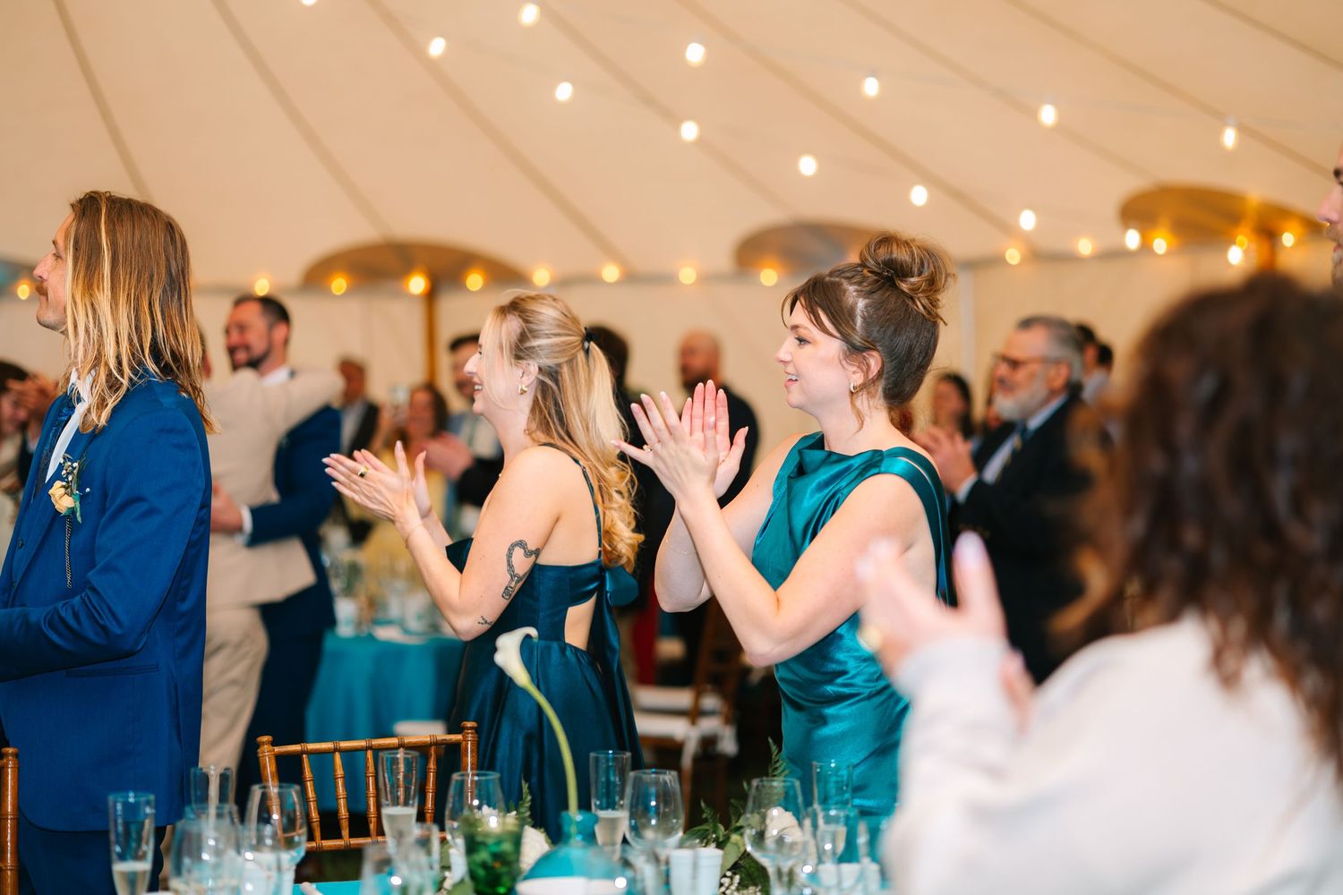 Wedding guests in teal and blue attire celebrate during a reception with string lights overhead.