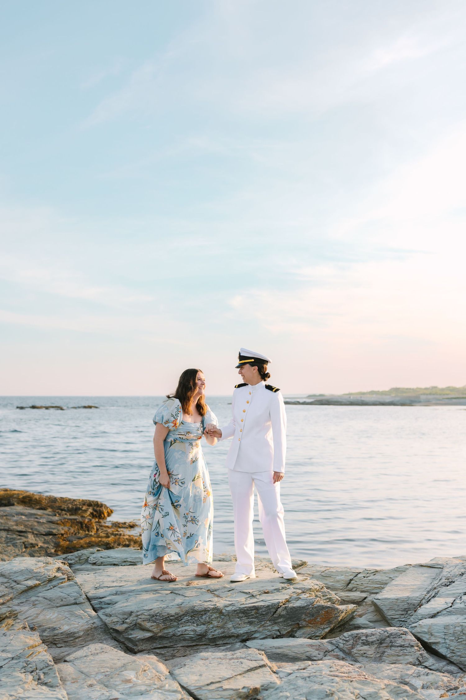 A couple holds hands and gazes at each other on rocky shoreline in naval uniform at dusk.