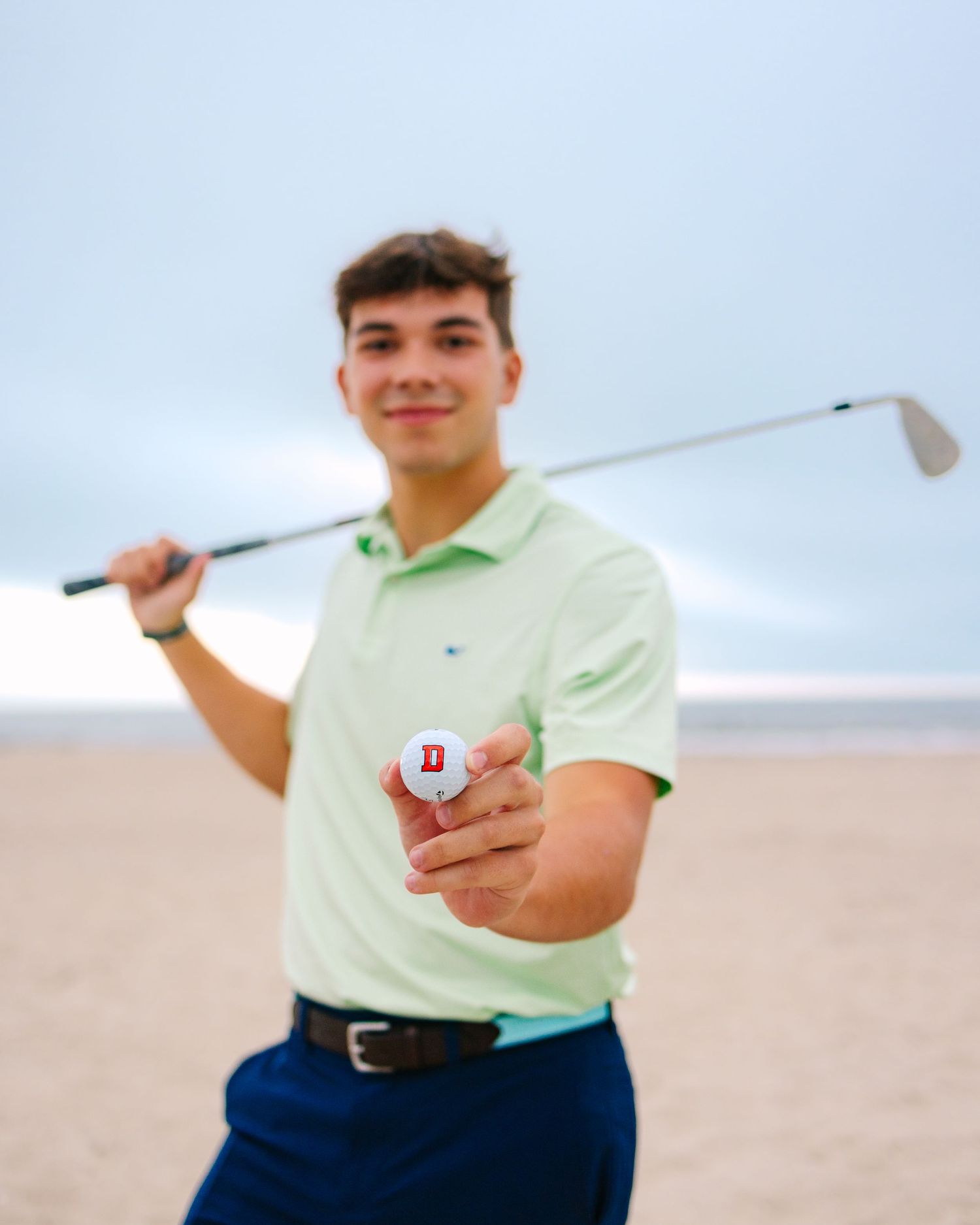 Young man in green polo holding golf club and ball on sandy beach.