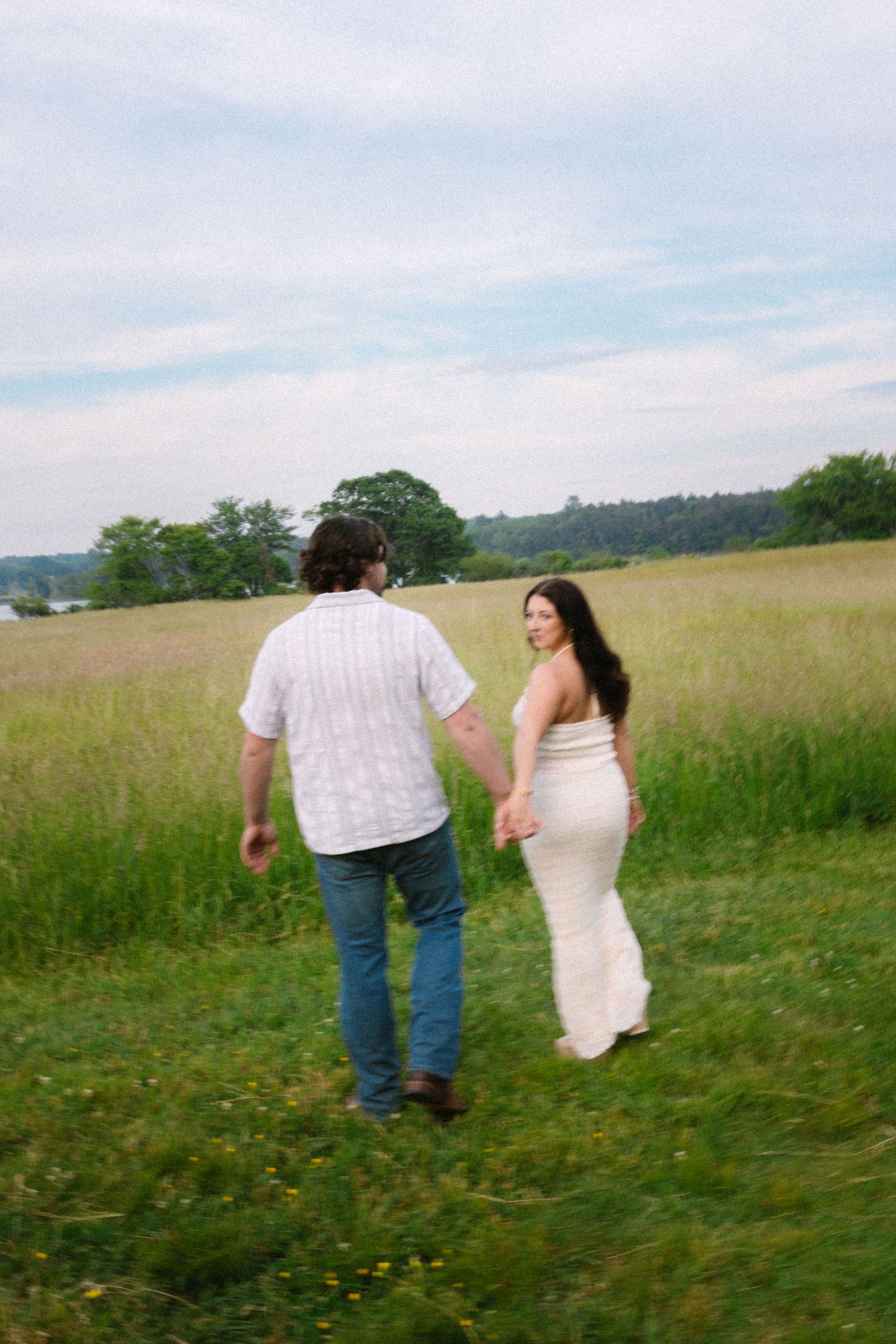 Couple walking away hand in hand through a lush green meadow toward distant trees.