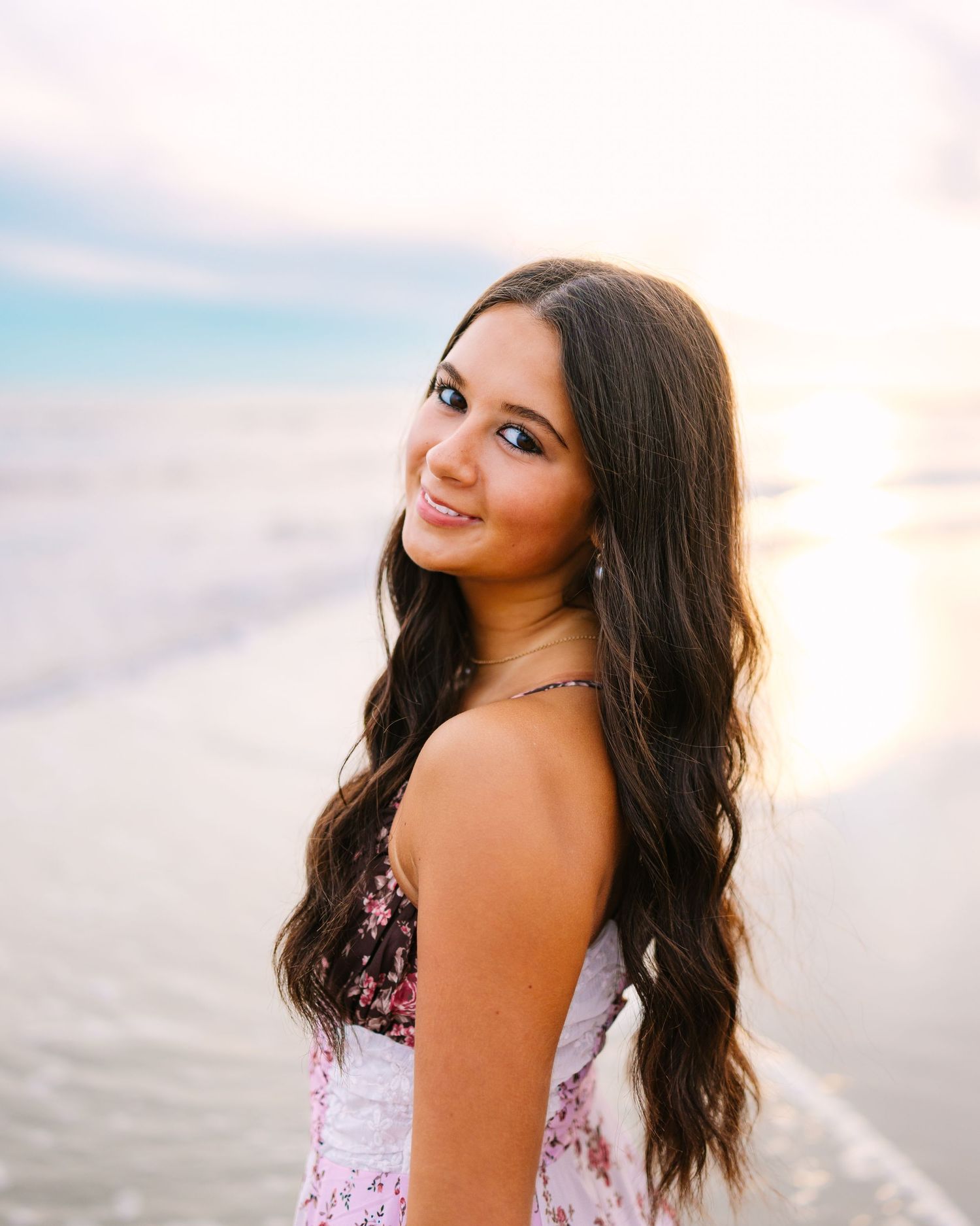 Smiling young woman with long wavy hair in a floral dress posing at a glowing beach sunset.