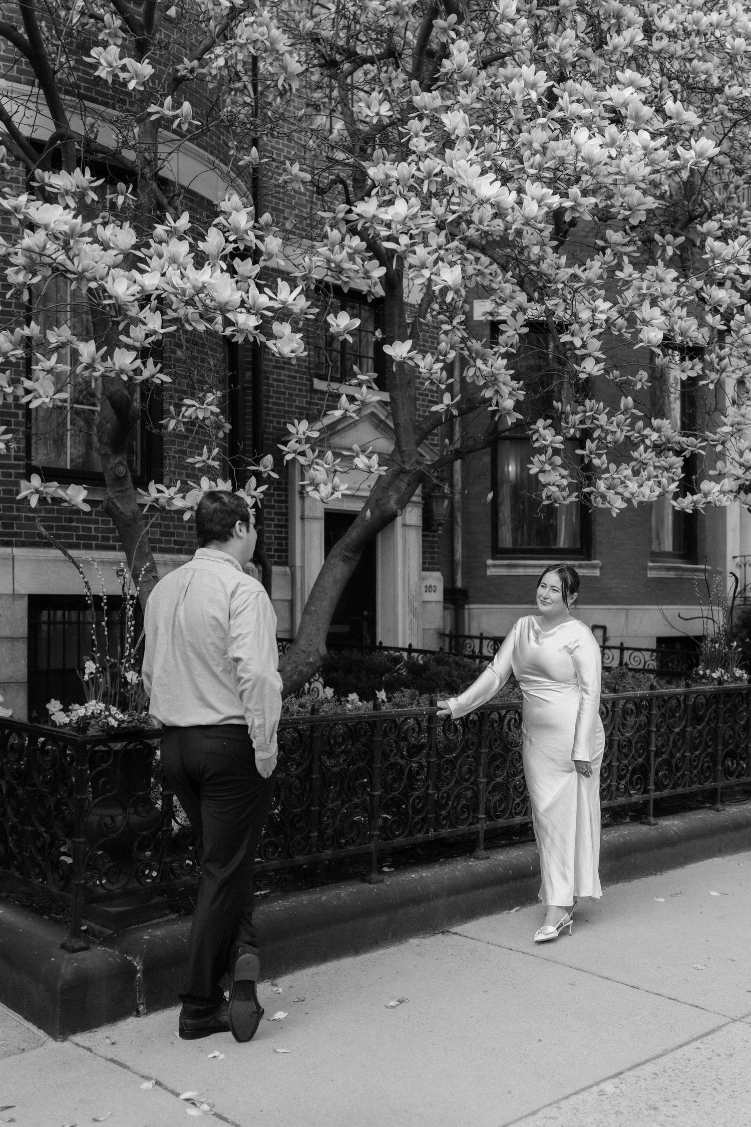 Black and white image of a couple holding hands beneath a blooming magnolia tree on a city sidewalk.