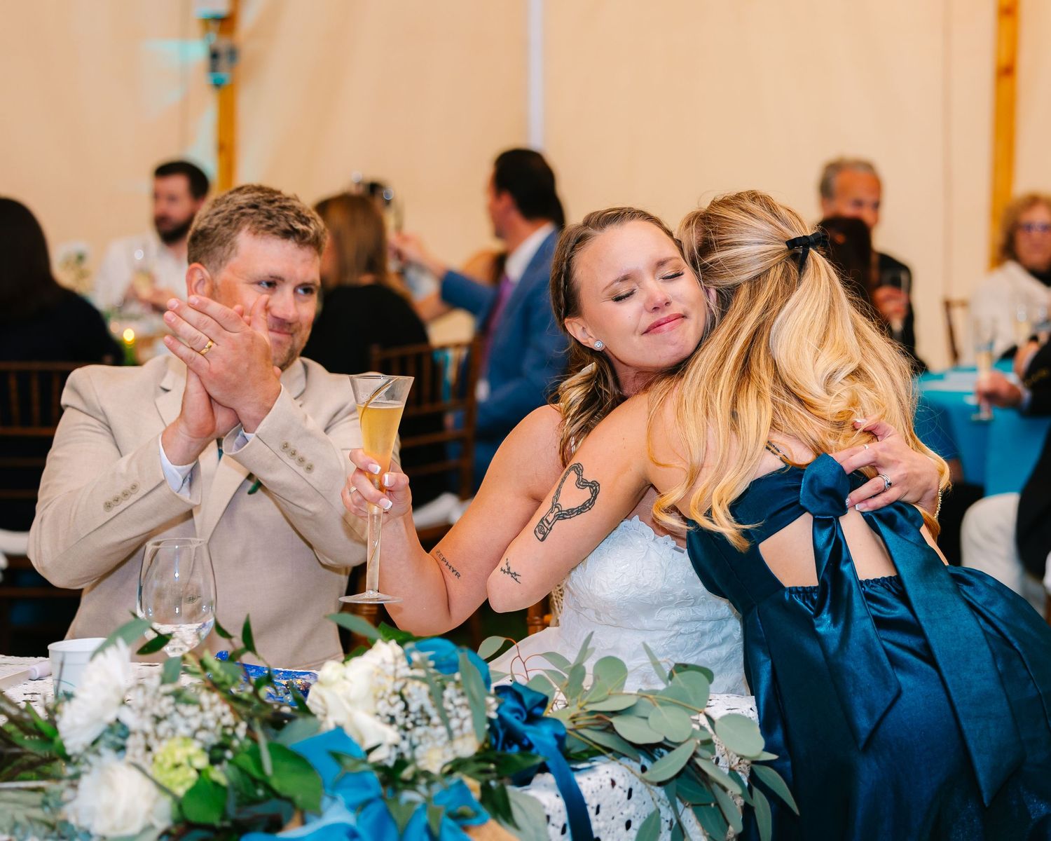 Wedding reception scene with emotional reactions at a table decorated with blue and green floral centerpieces at backyard tented reception