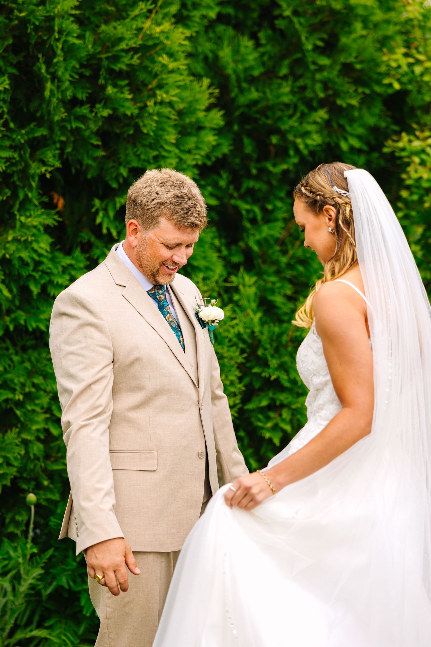 A romantic moment between a bride in white and groom in beige captured in a lush green garden setting.
