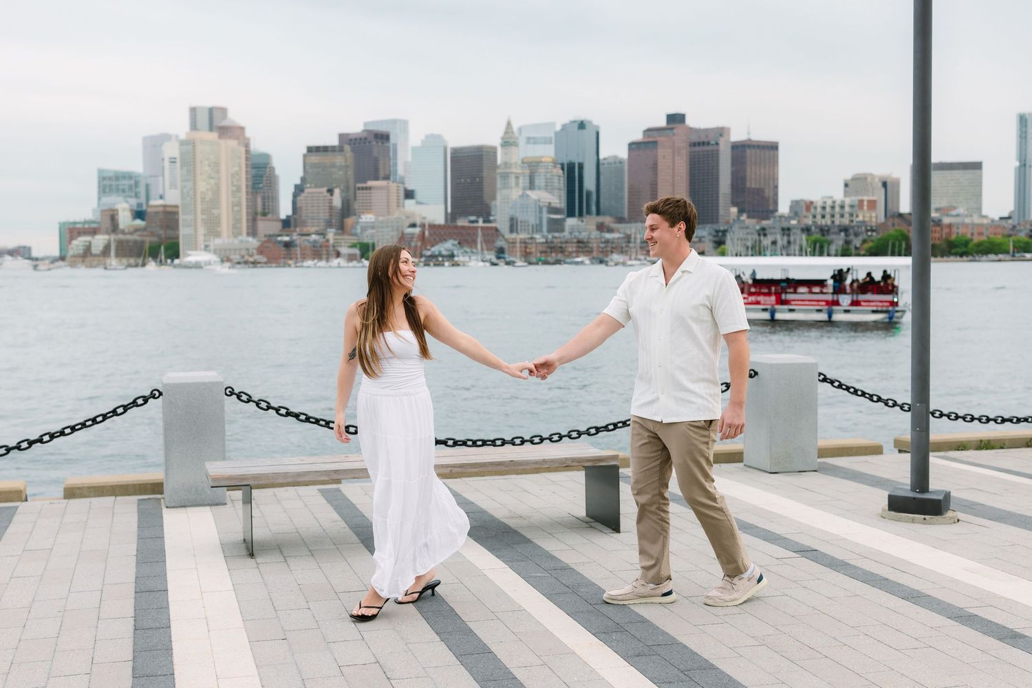Engaged couple holding hands on a Boston waterfront pier with city skyline backdrop.
