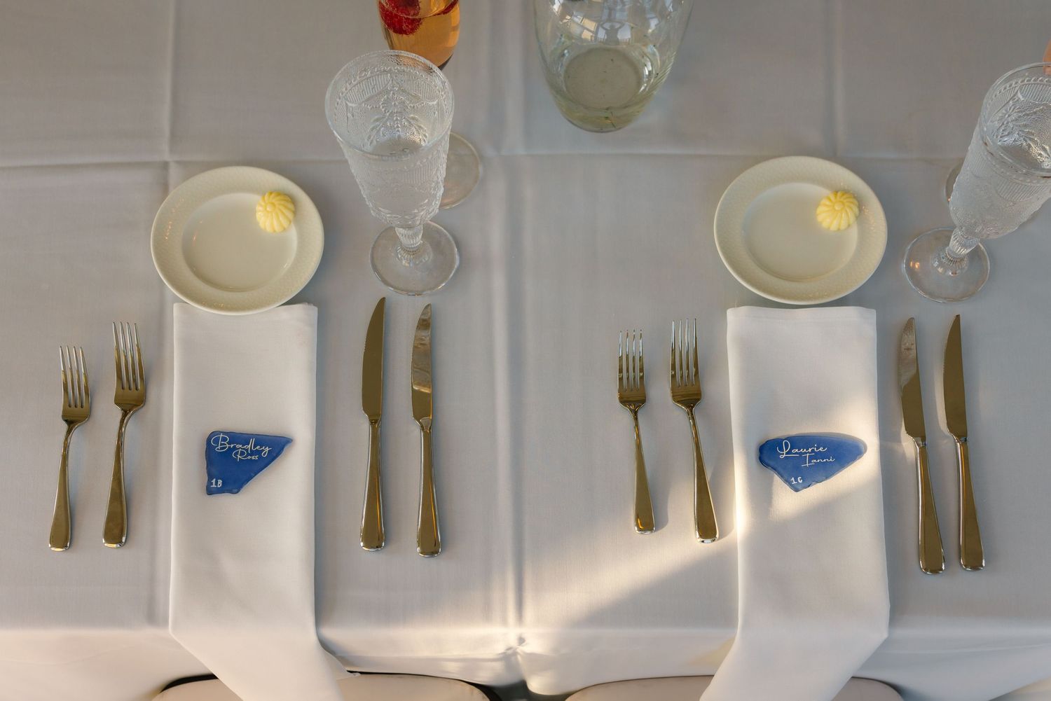 Elegant wedding reception table setting with gold cutlery, white napkins, crystal glasses, and blue place cards on white tablecloth.