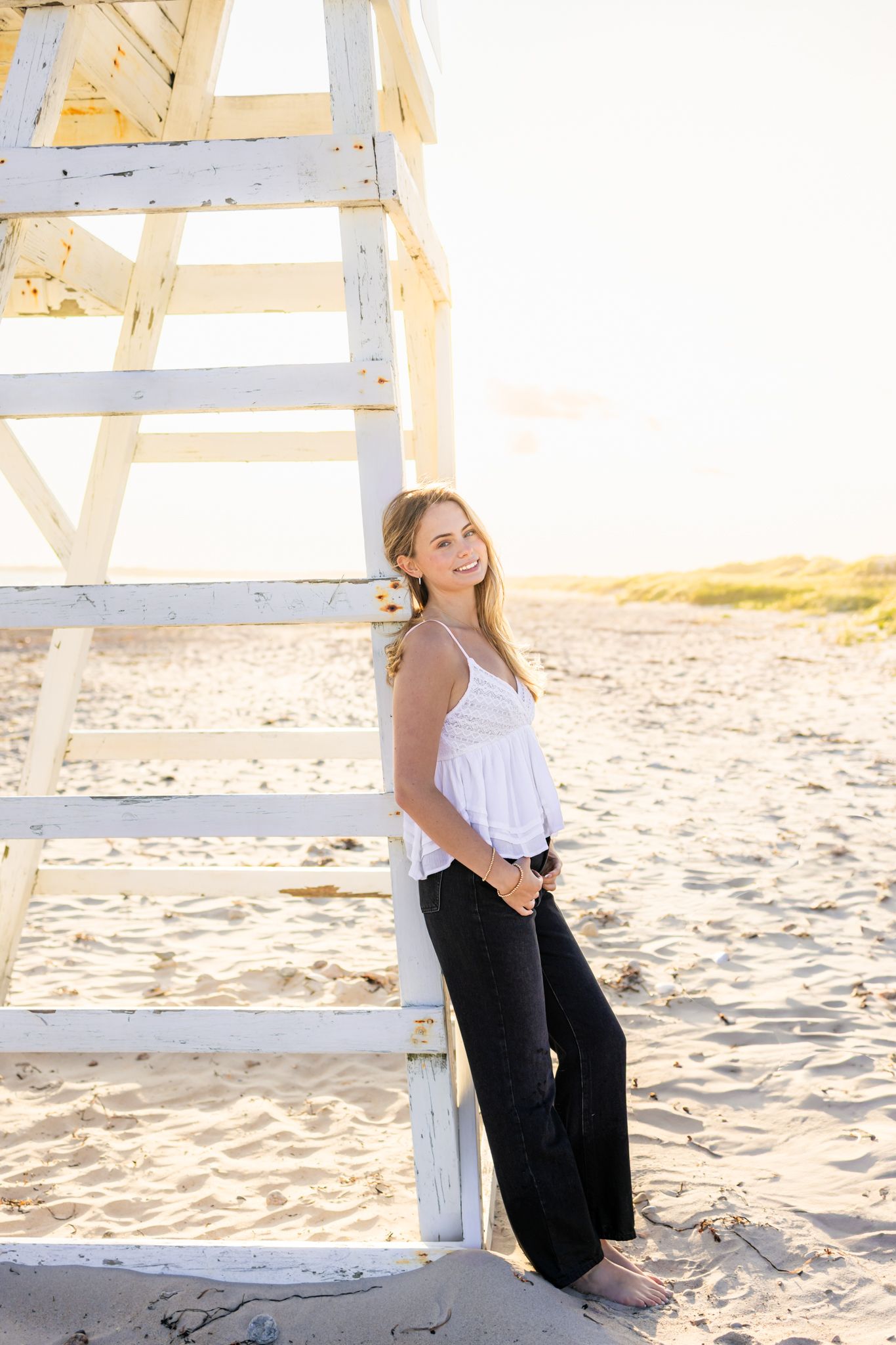 Young woman in white top and black pants leaning against a lifeguard stand on a sunny beach.