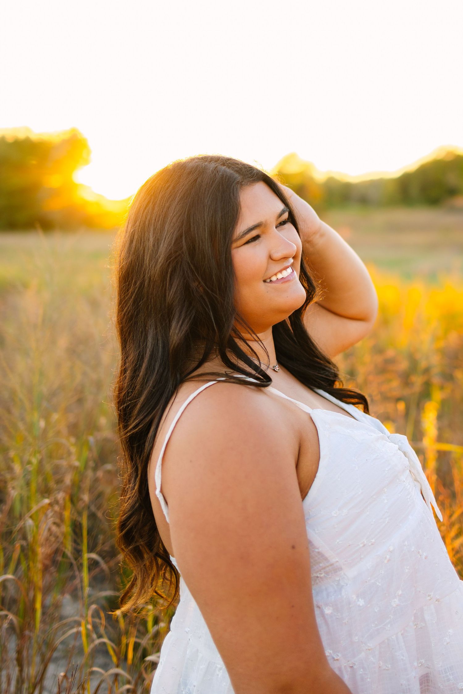 Smiling young woman in white dress with hand in hair in golden sunlit field.