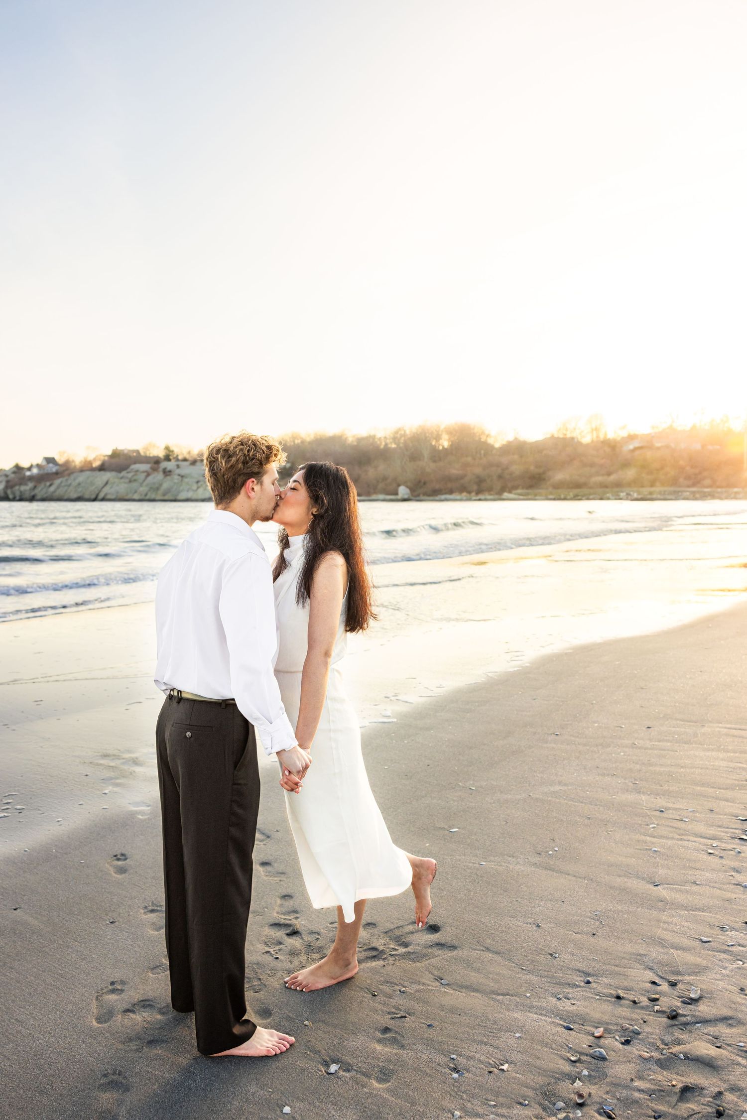 A couple shares a romantic moment at sunset on a sandy beach while dressed in elegant white and black attire.