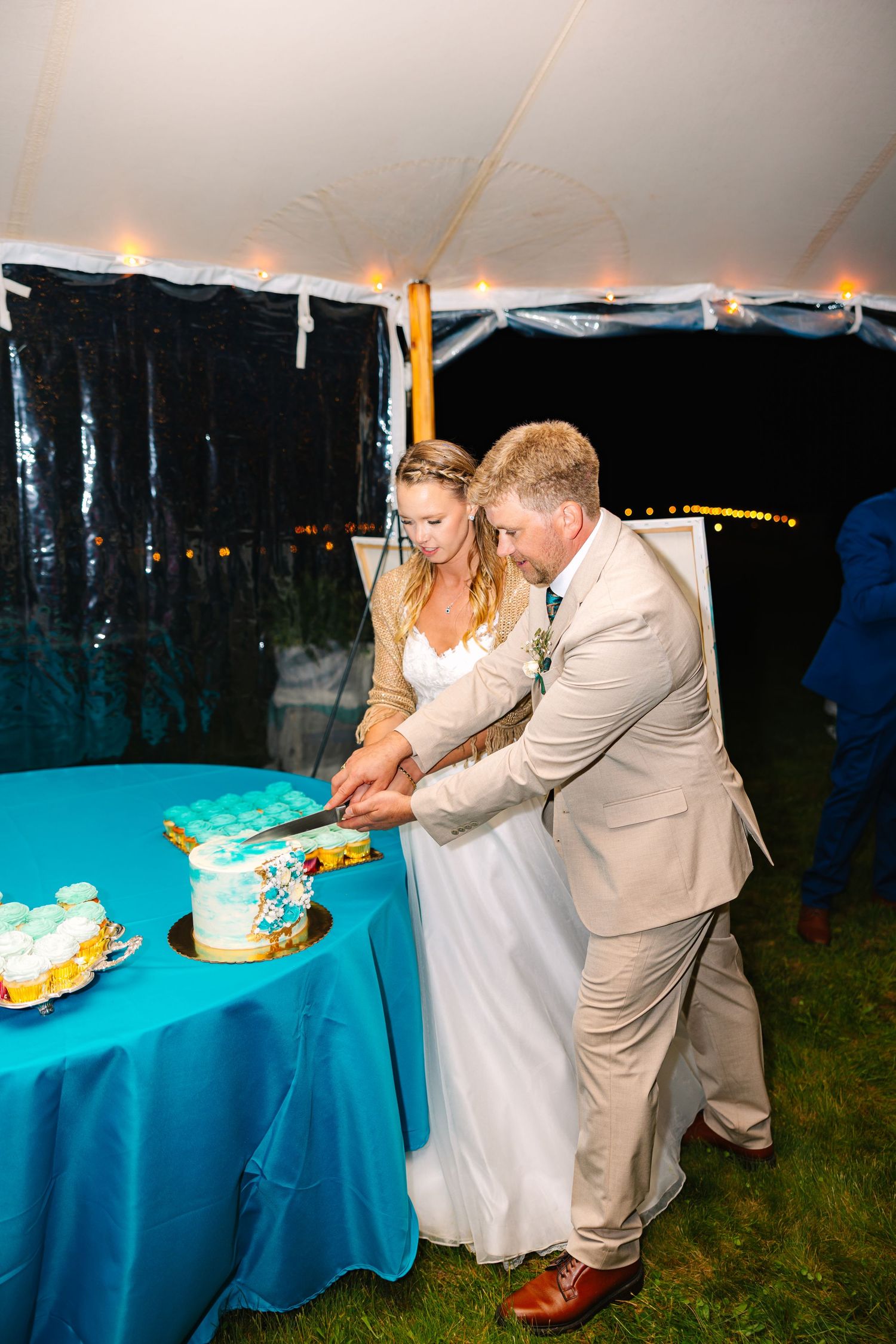 A couple in wedding attire cuts their blue and white wedding cake together at an outdoor reception with turquoise decor.