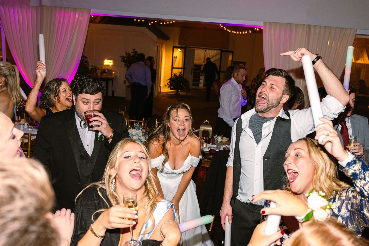 Energetic crowd dancing and celebrating at a wedding reception with pink decor and uplighting.