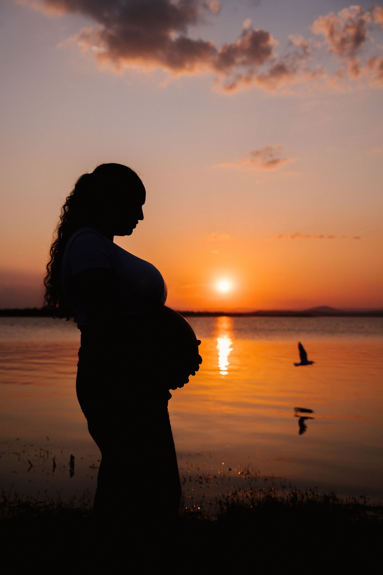 Silhouette of pregnant woman cradling baby bump against vibrant orange sunset reflecting on calm lake.