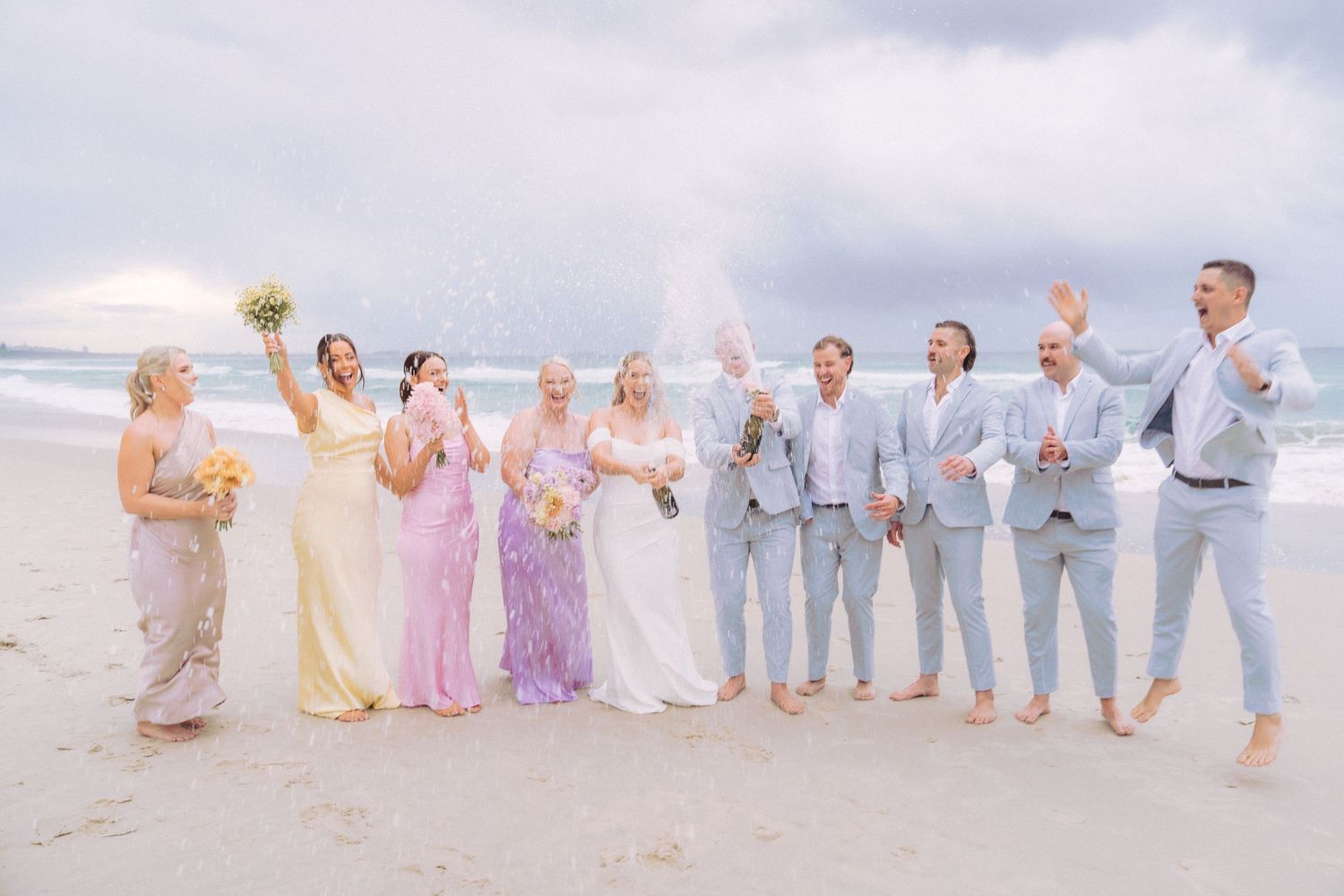Bride and bridal party celebrating with a champagne spray on the beach after the wedding ceremony.
