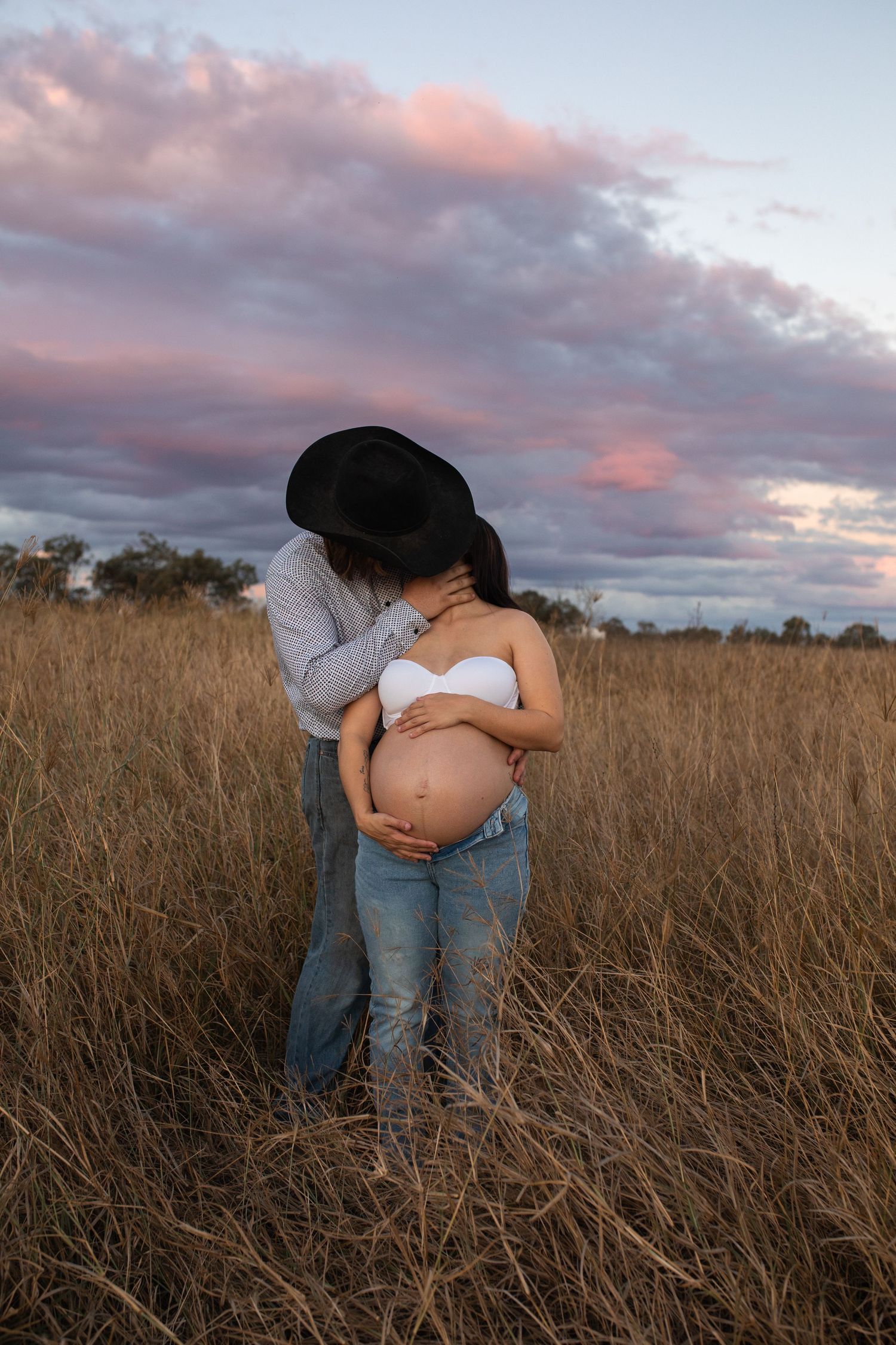 Couple embracing in golden field at sunset, pregnant woman in white bralette and jeans, man in cowboy hat.