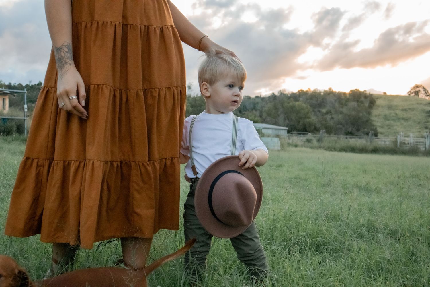 Young toddler boy holding hat standing beside mother in rust orange dress in rural field.