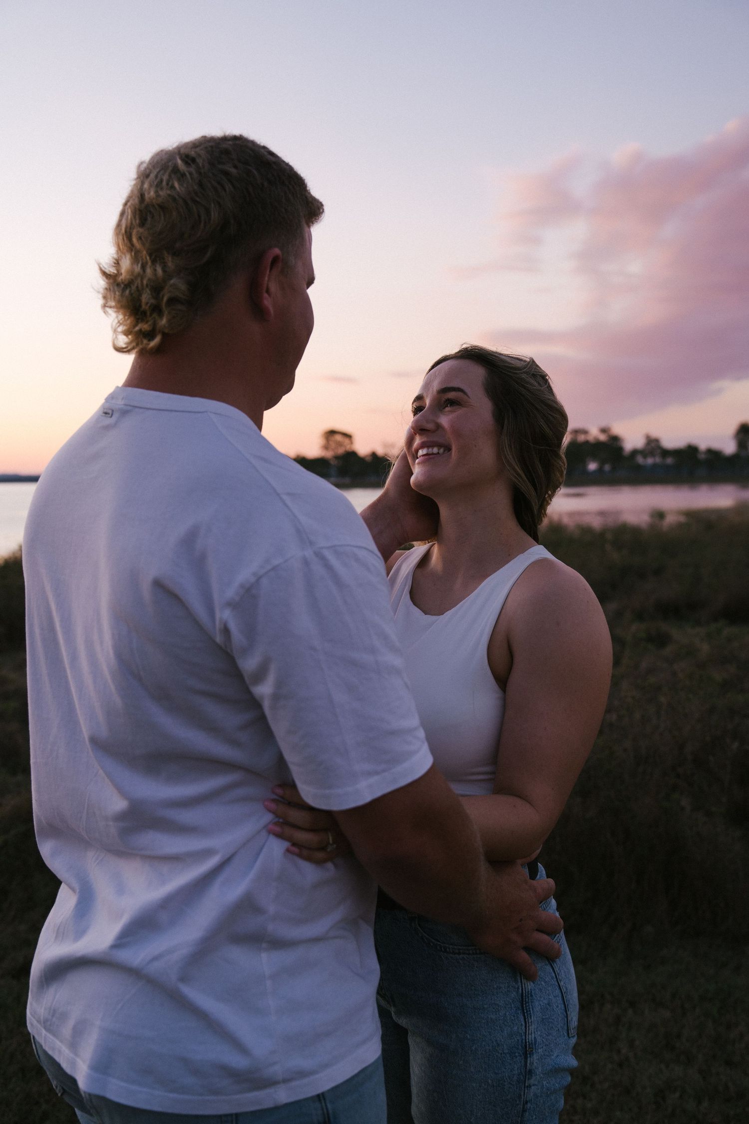 Couple embracing at sunset near a lake, woman smiling up at man, both wearing white tops in a romantic outdoor setting.
