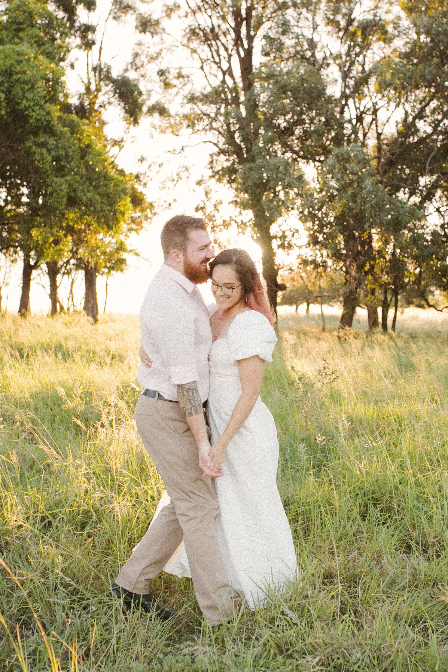 Couple holding hands and walking through a sunlit grassy field with trees at golden hour.