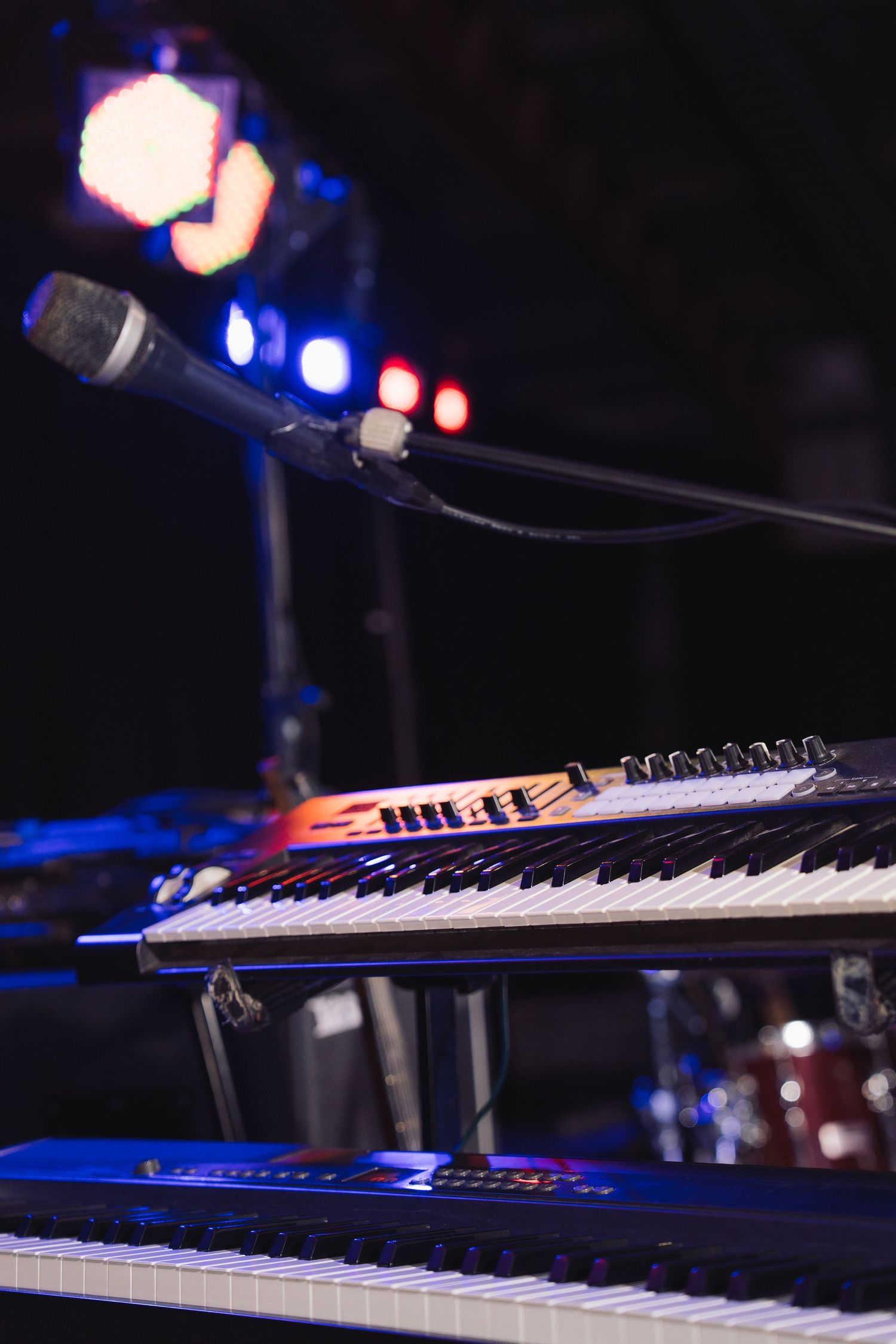 Two keyboards on a concert stage with a microphone stand and colorful stage lights in the background.