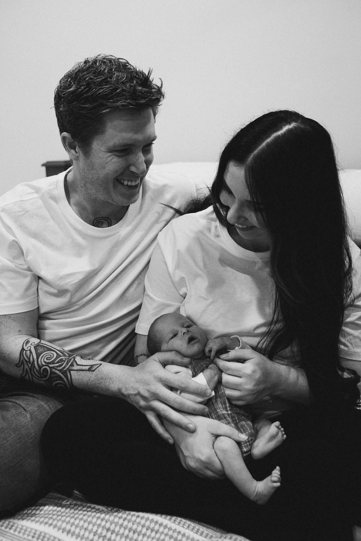 Black and white photo of smiling parents with tattoos cradling their newborn baby on a bed.