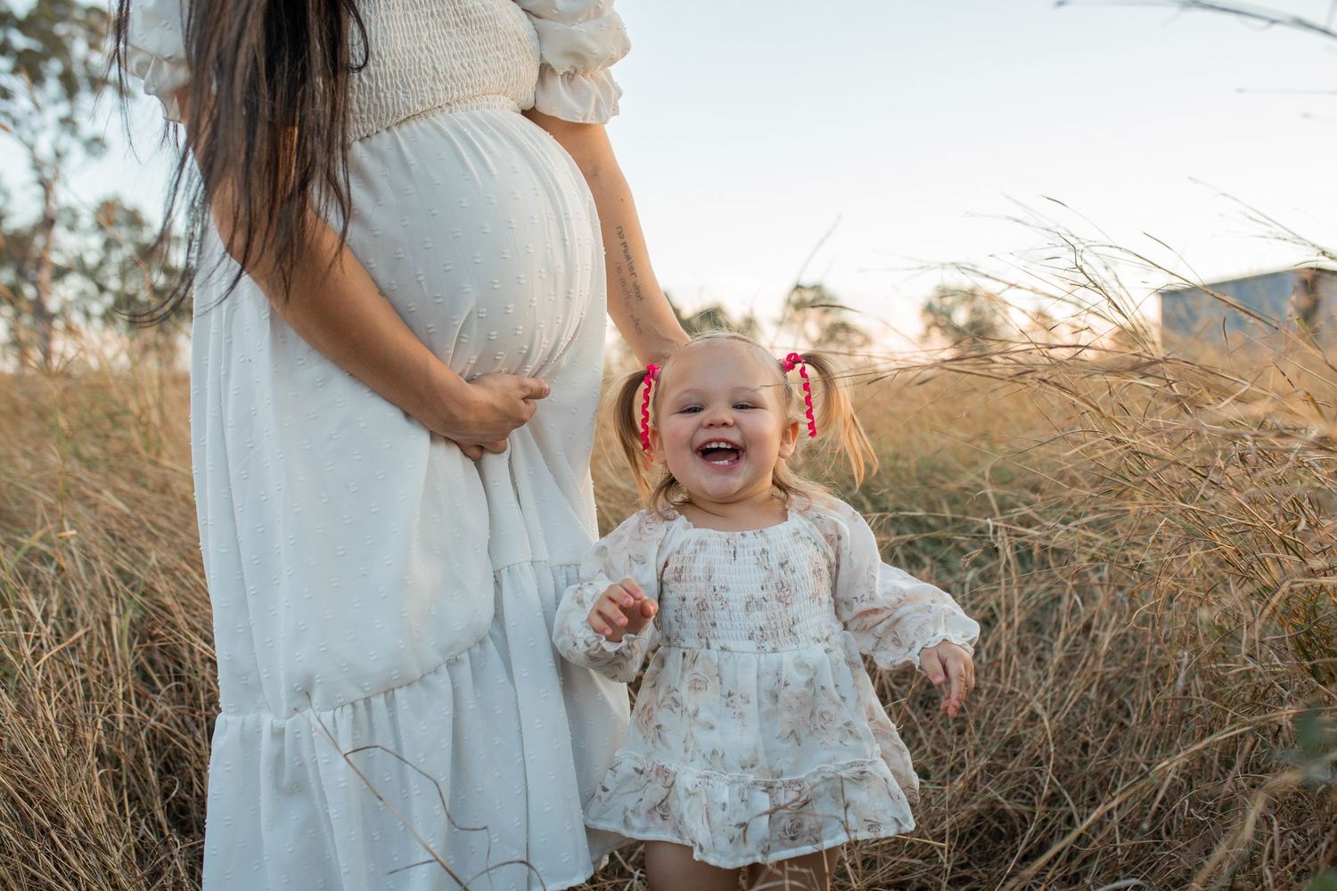 Pregnant woman in white dress standing in golden field with laughing toddler girl wearing floral dress and red hair bows.