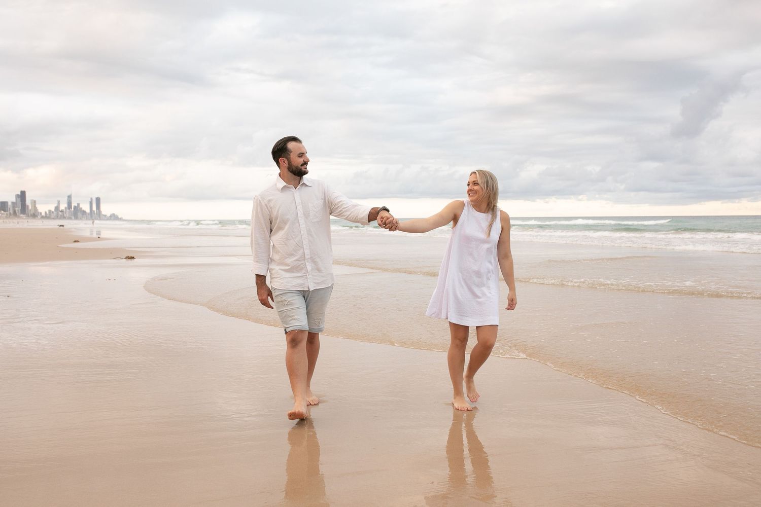 Couple holding hands on the shoreline during a relaxed beach engagement session.
