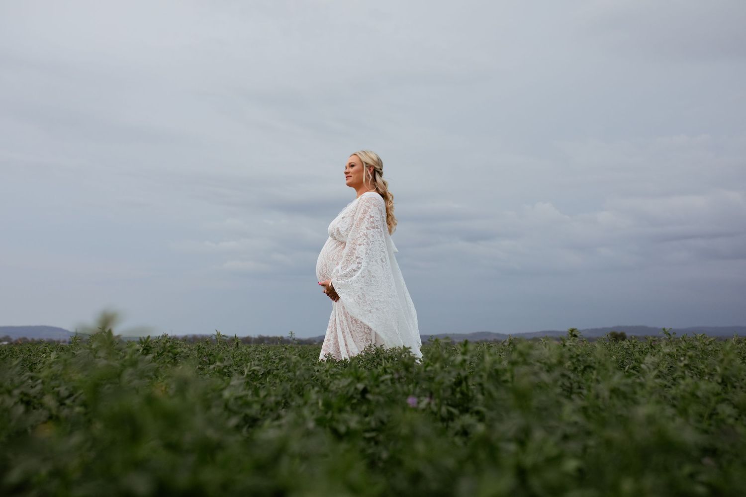 Pregnant woman in white lace dress walking through lush green field on overcast day.