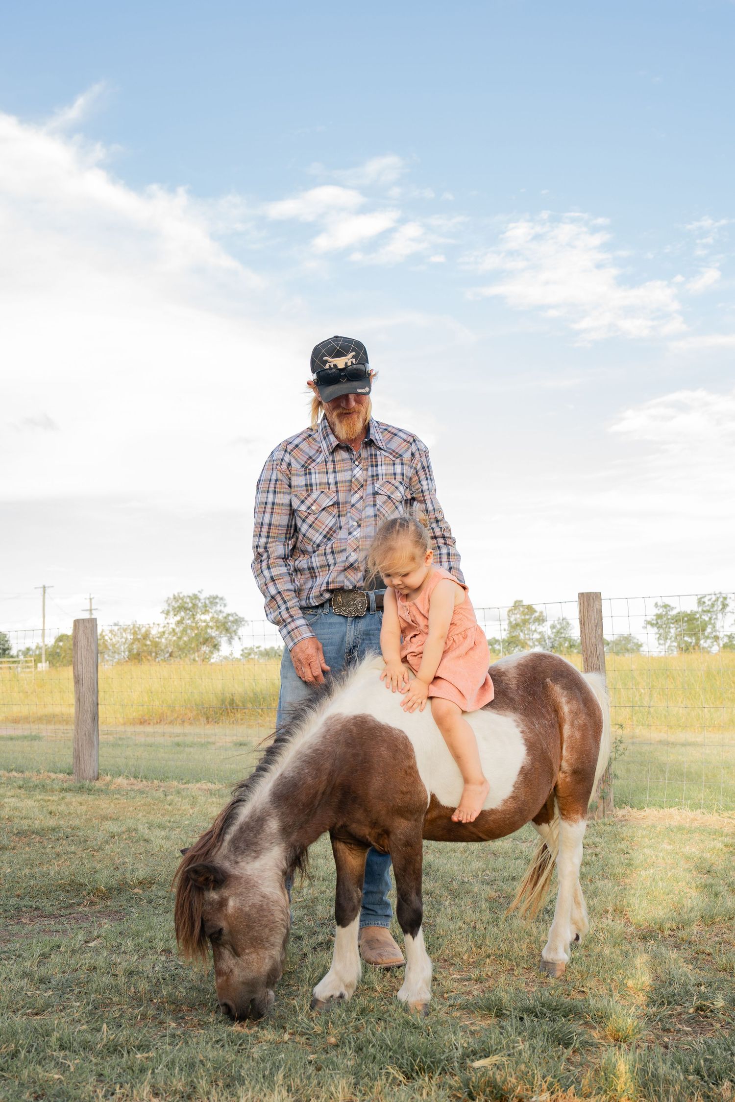 An elderly man in a plaid shirt and cap helps a young girl ride a brown and white pony on a sunny farm.