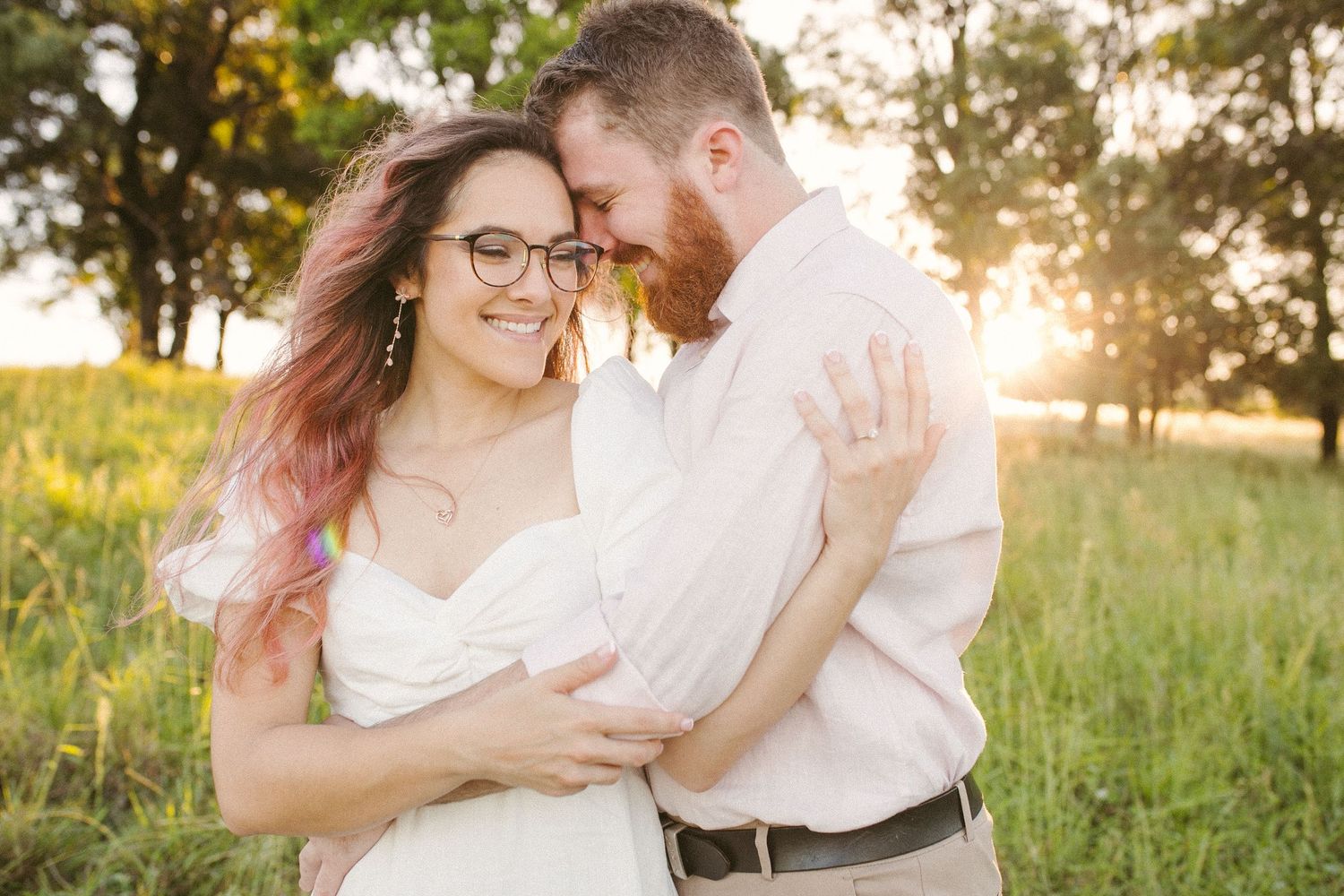 Smiling couple embracing in a sunlit meadow with golden hour light glowing behind them.