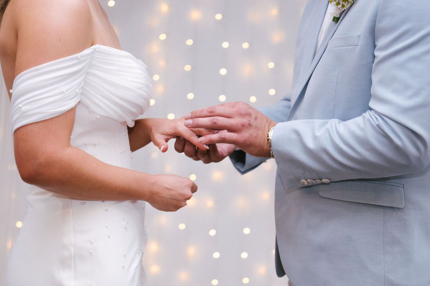 Bride and groom exchanging wedding rings beneath glowing fairy lights during the ceremony.