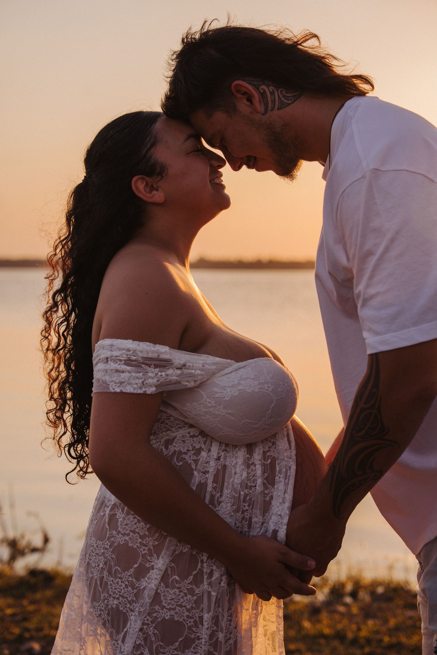 Pregnant couple sharing a romantic moment at sunset by the water, woman in white lace dress.