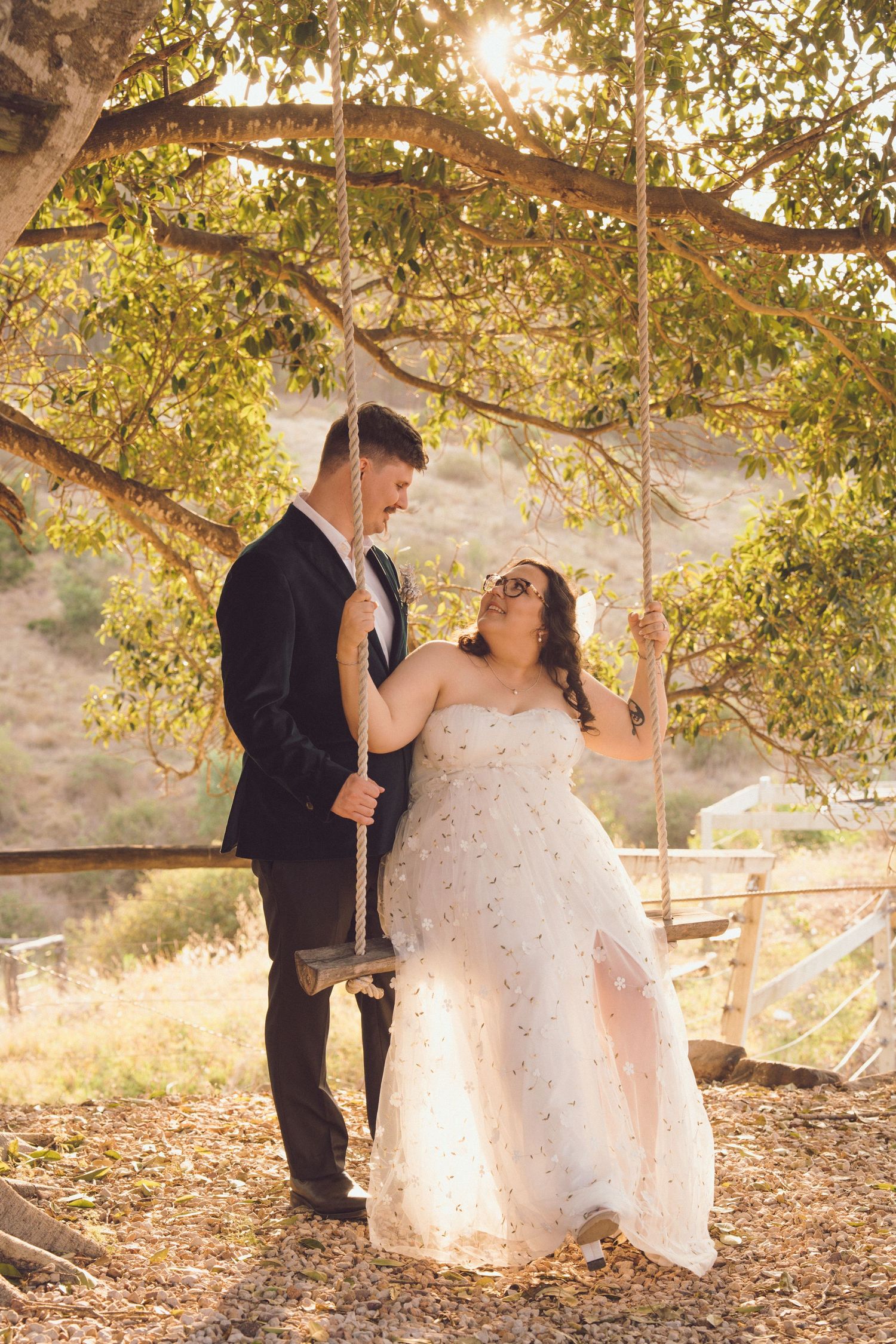 Bride sitting on a swing while smiling up at the groom in warm golden evening light.