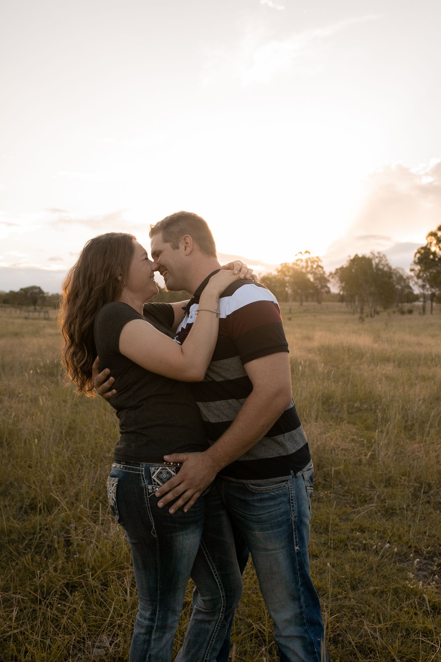 Couple embracing and almost kissing in a golden sunset field, wearing casual jeans and t-shirts in a romantic outdoor setting.