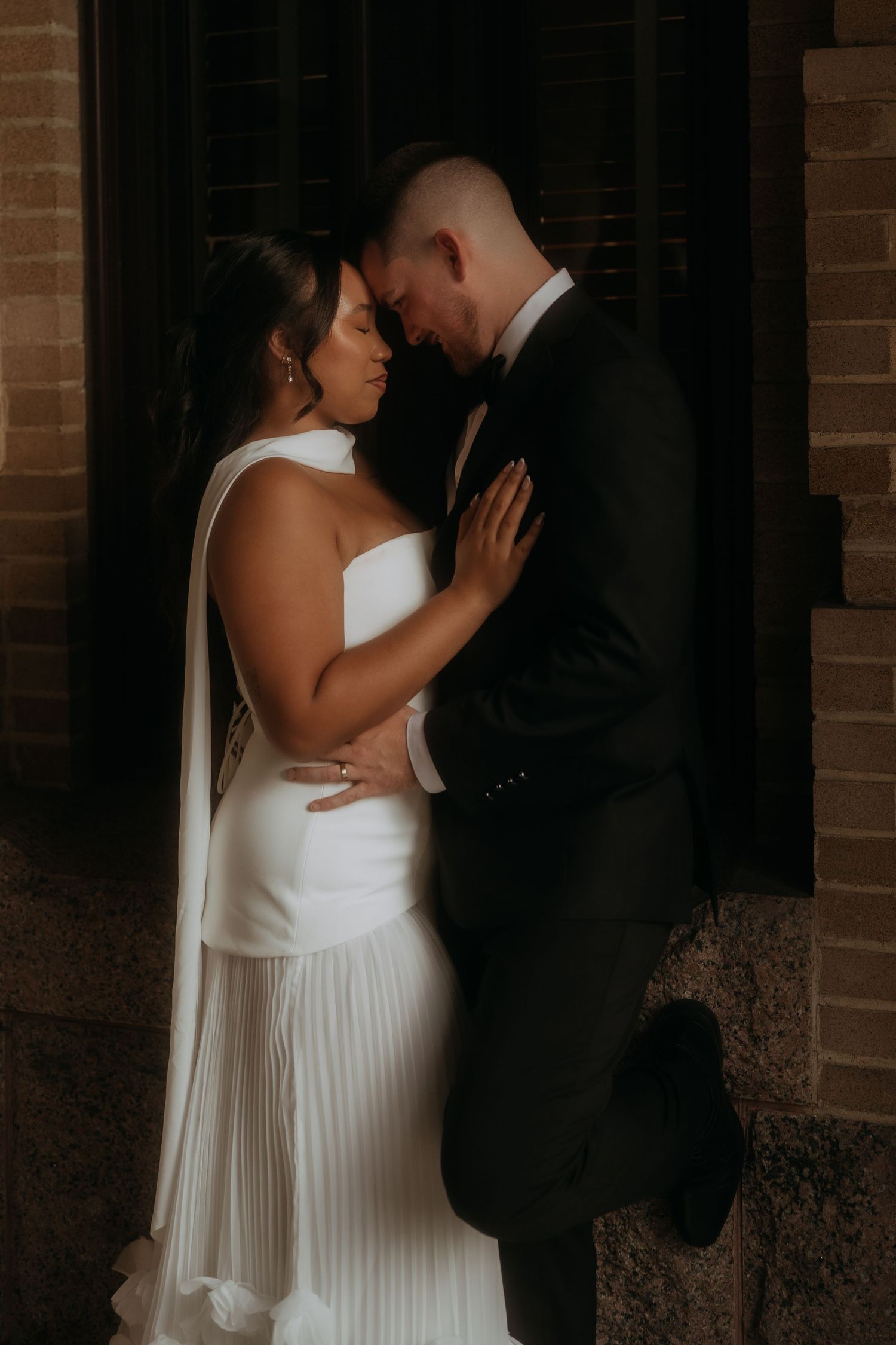 Romantic wedding couple touching foreheads intimately in dramatic shadow against brick wall.
