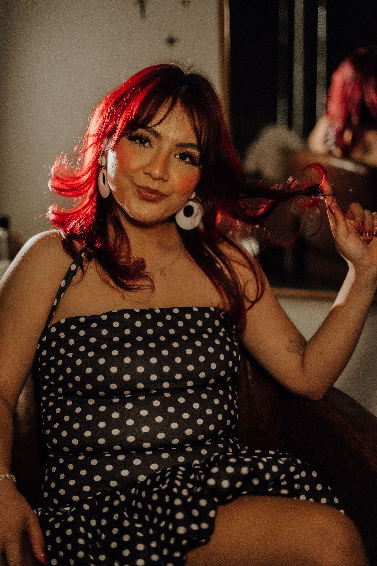 Woman with vibrant red hair in black polka dot dress sitting in salon chair holding her hair in warm light.