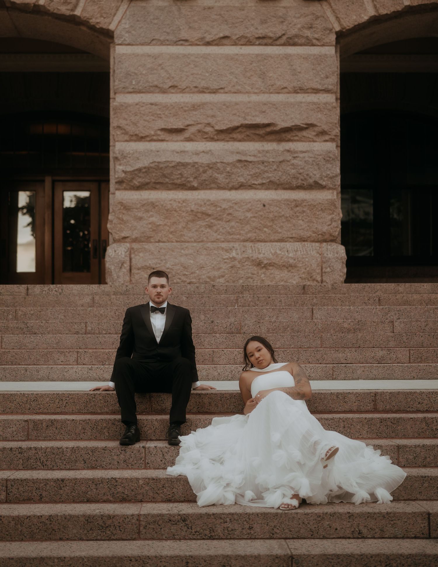 Bride in white gown and groom in black tuxedo sitting on granite steps of historic building.